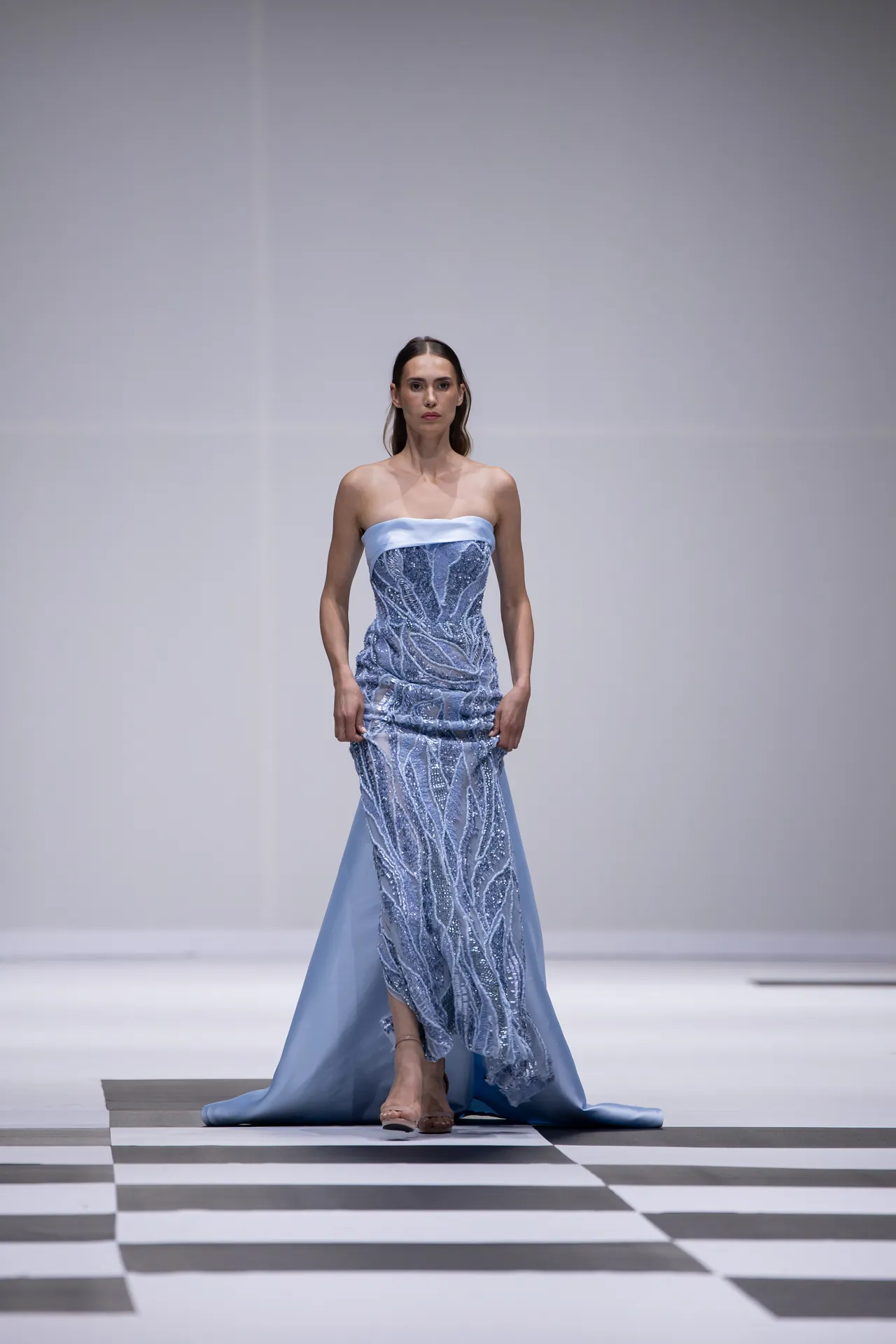 Model on a runway wearing a strapless light-blue gown with silver bead embroidery, walking down a checkered catwalk.