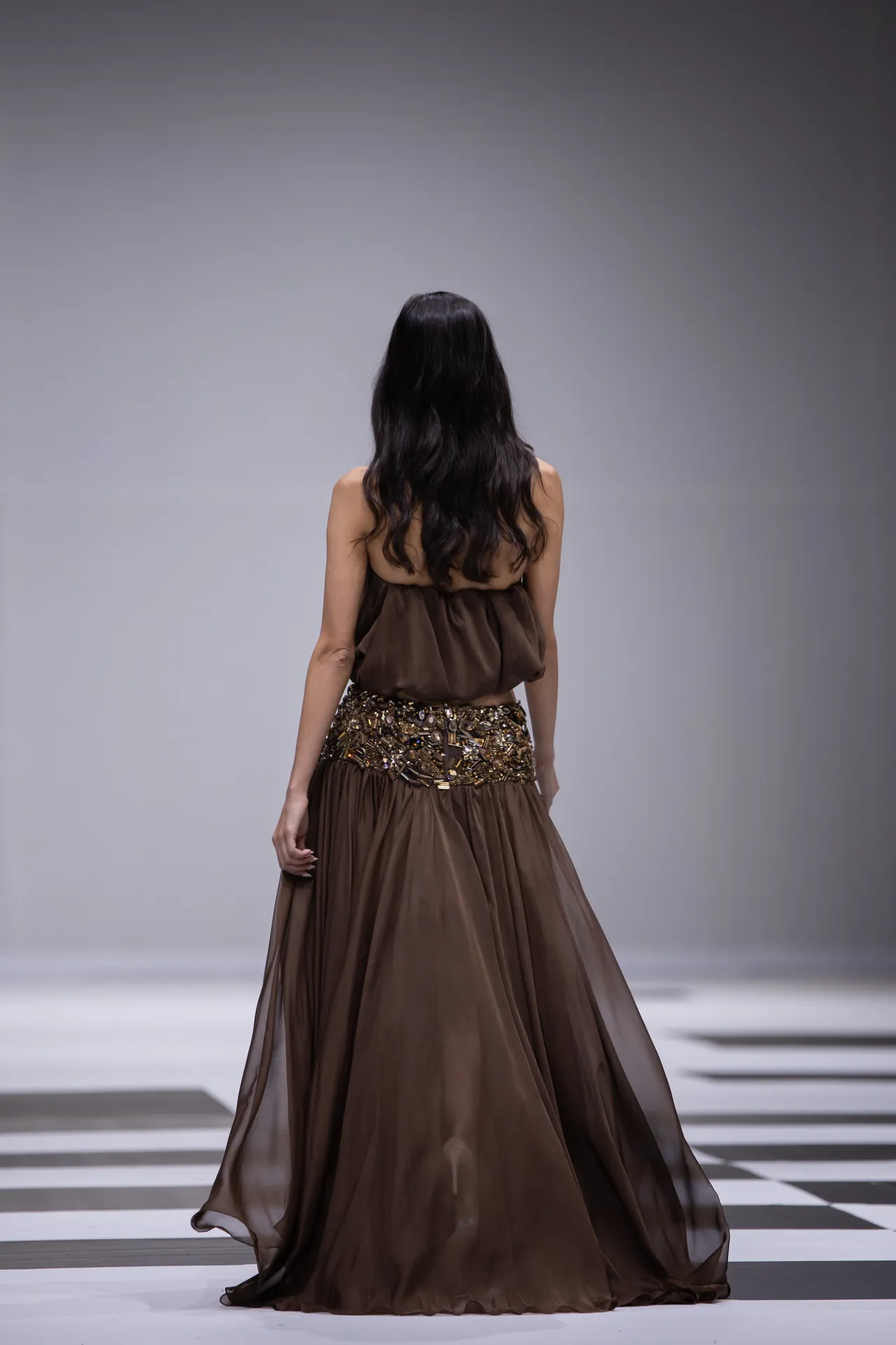 Back view of a model wearing a floor-length brown gown with a jeweled belt walking on a checkered runway.
