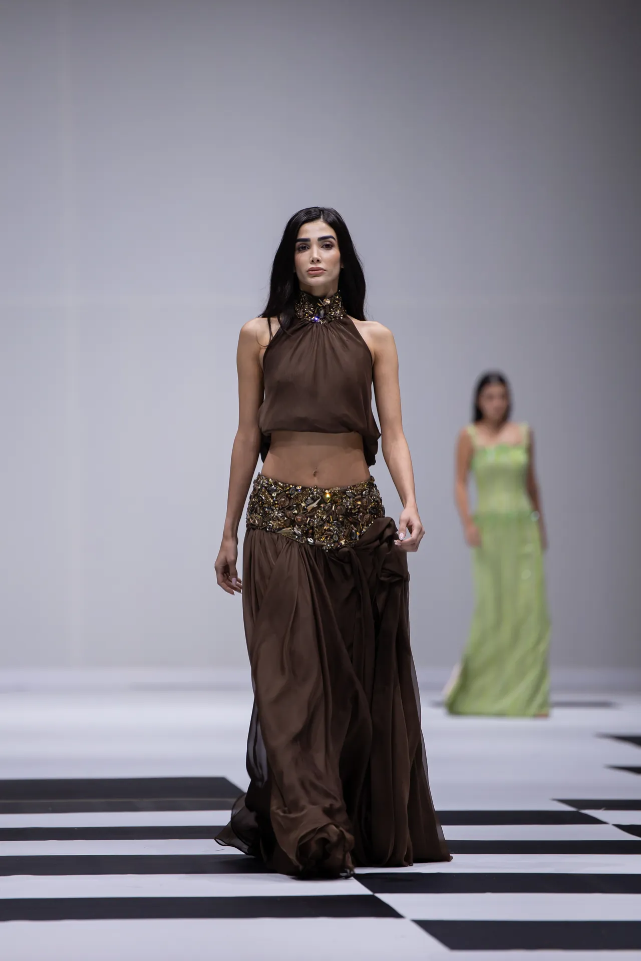 Model in a brown halter draped gown with a jeweled belt walks a runway on a black-and-white checkered floor; a second model in a lime dress appears blurred in the background.