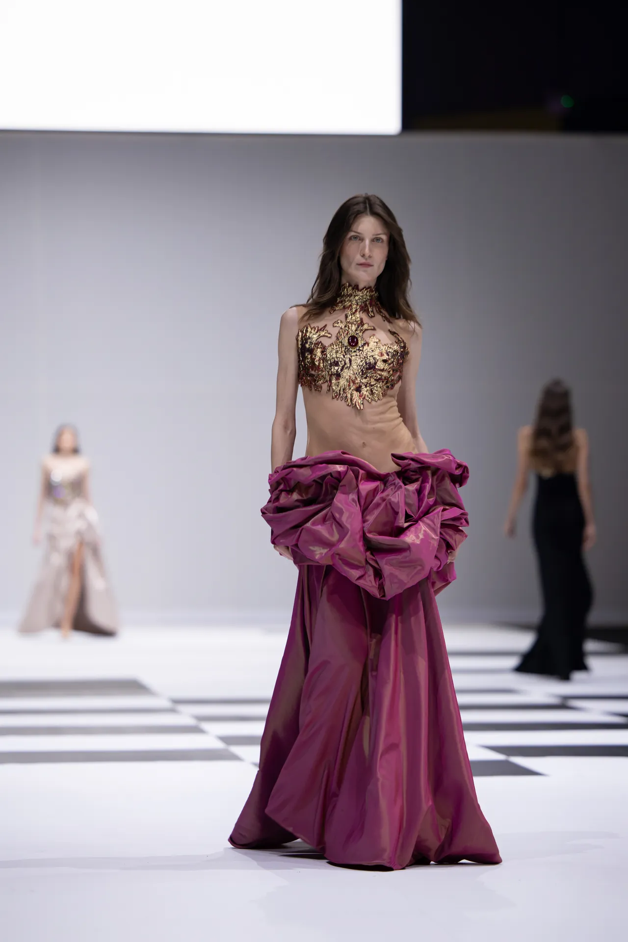 Model walks the runway in a gold-embellished bodice and a dramatic magenta skirt, with a checkered floor backdrop.