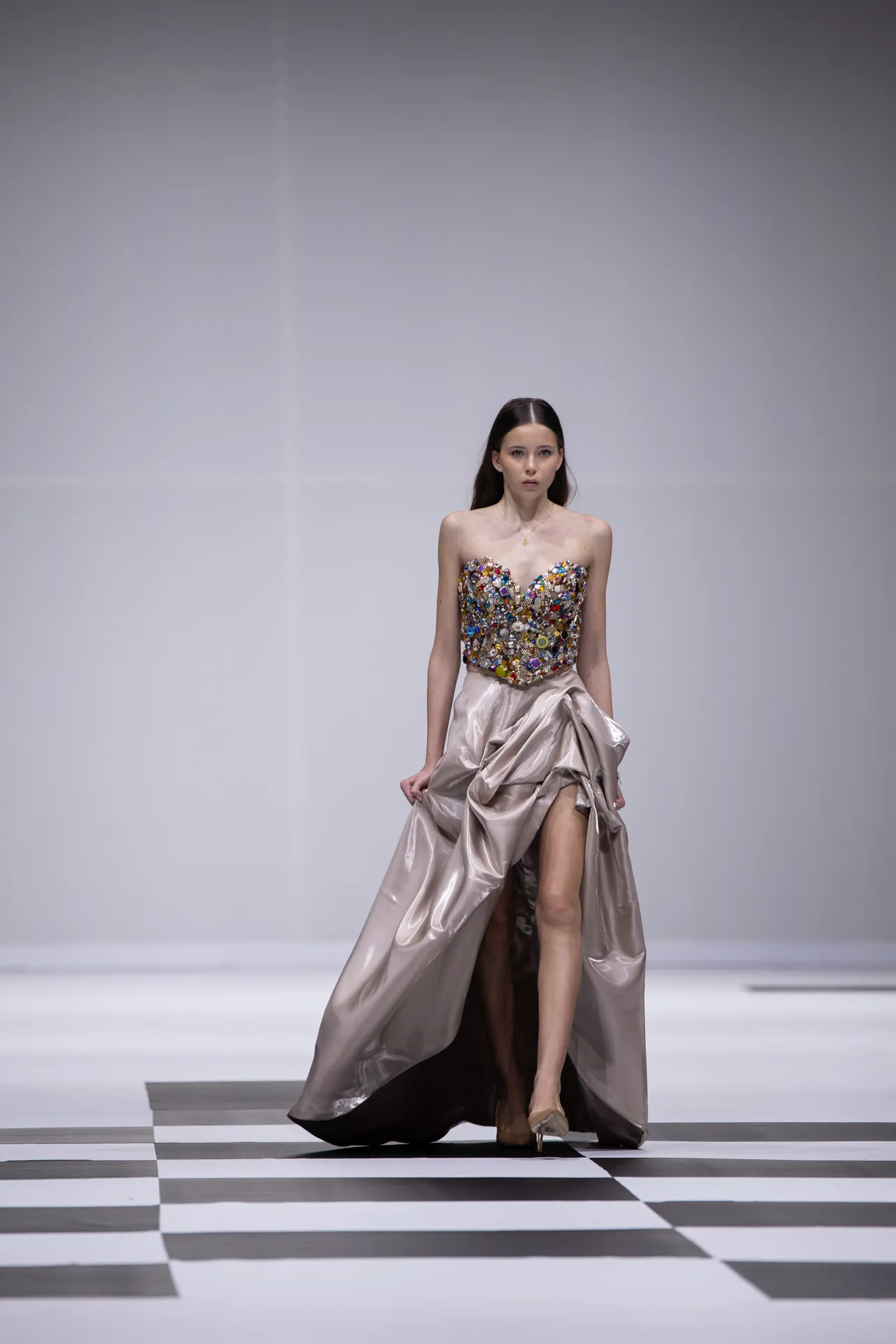 Model walks the runway in a strapless, jewel‑embellished bodice gown with a satin, flowing skirt on a black‑and‑white checkered floor.