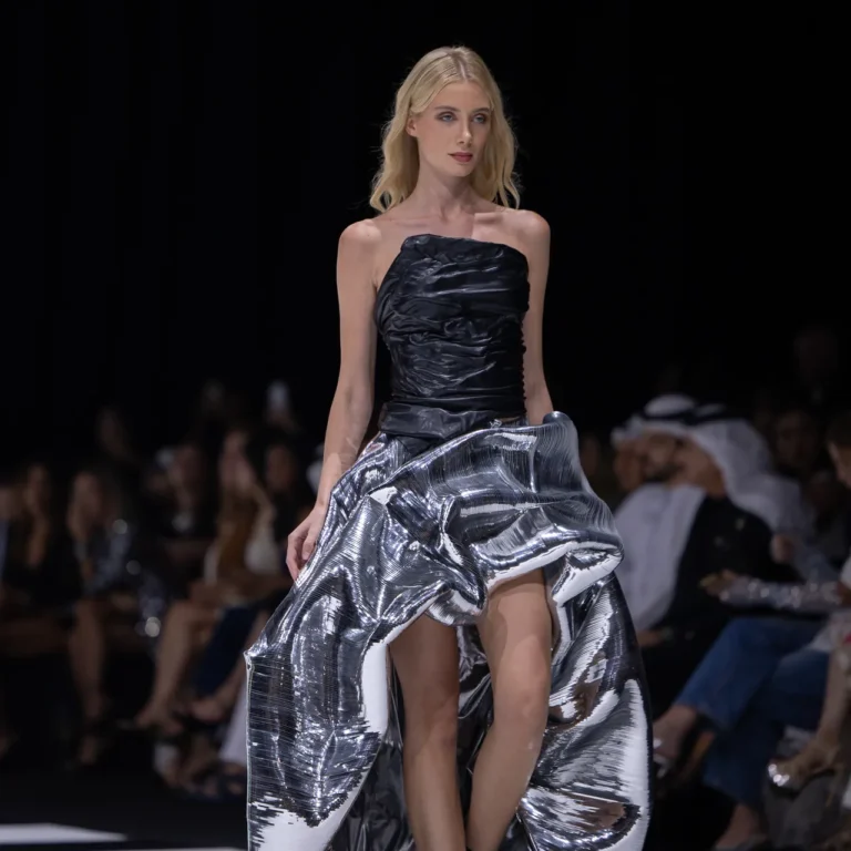 Model walks the runway in a strapless black bodice and a shiny, voluminous metallic silver skirt, mid-stride as the audience watches.
