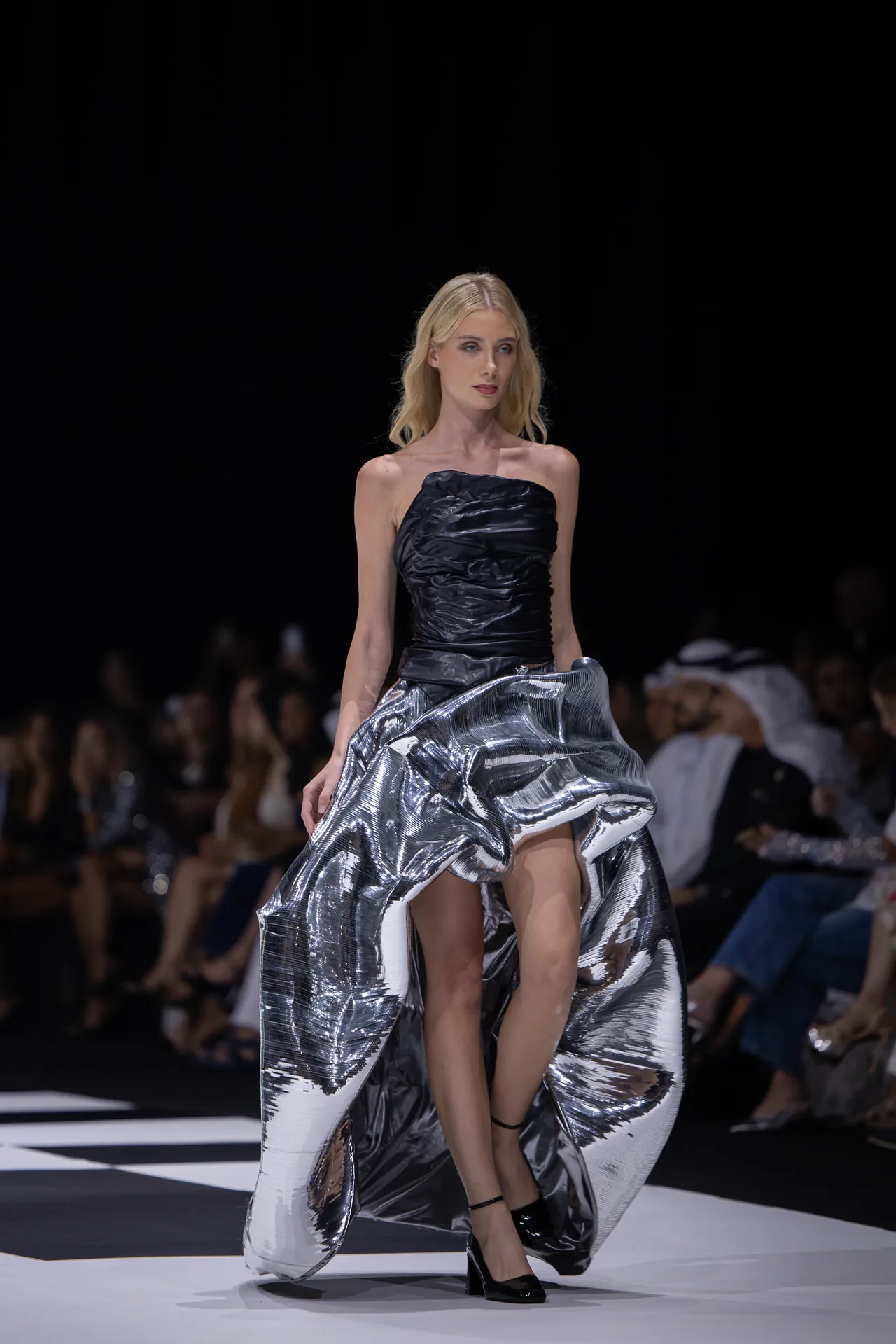 Model walks the runway in a strapless black bodice and a shiny, voluminous metallic silver skirt, mid-stride as the audience watches.
