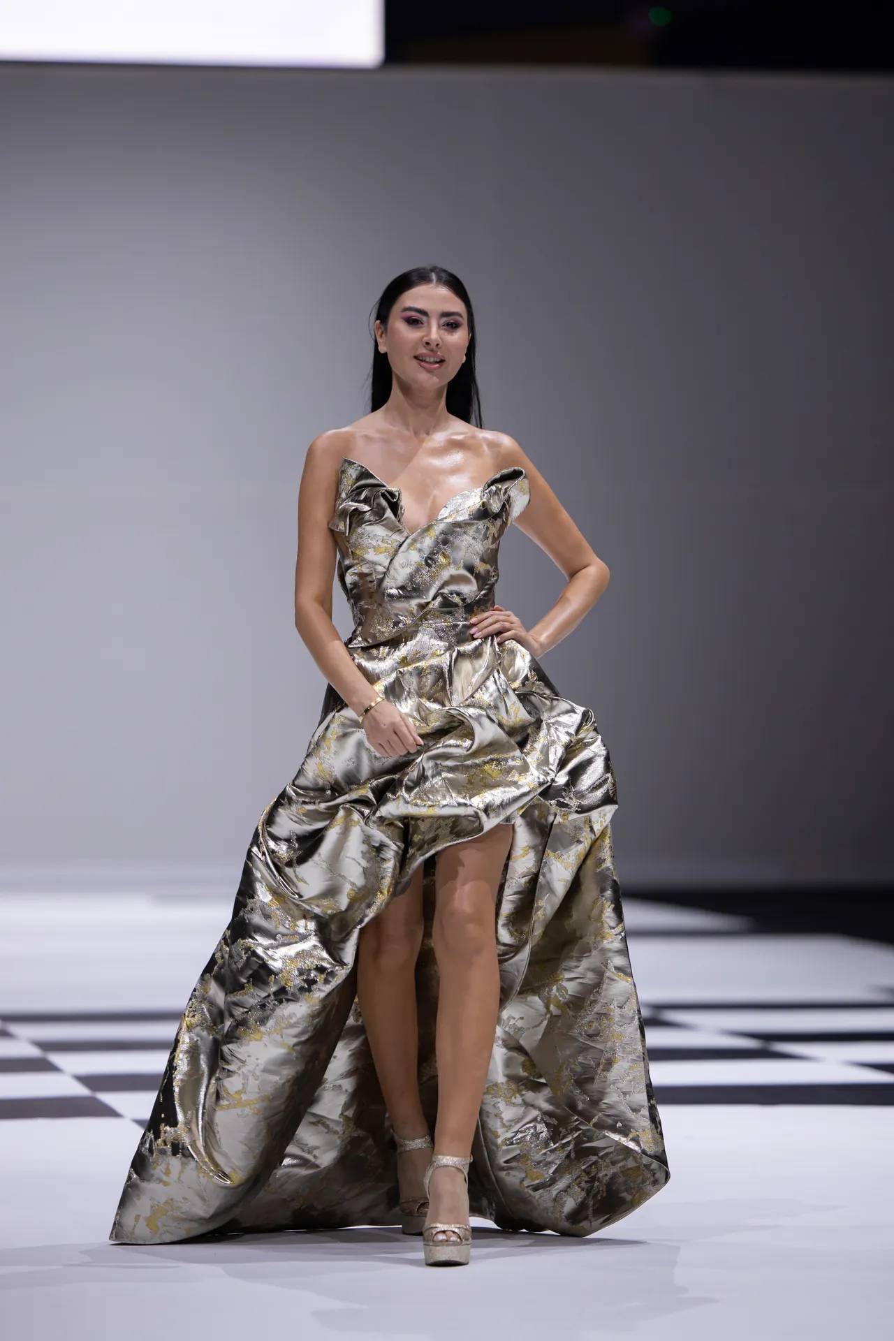 Fashion model walking a runway in a metallic gold-silver floral gown with large ruffle bodice and voluminous train, checkered floor background.
