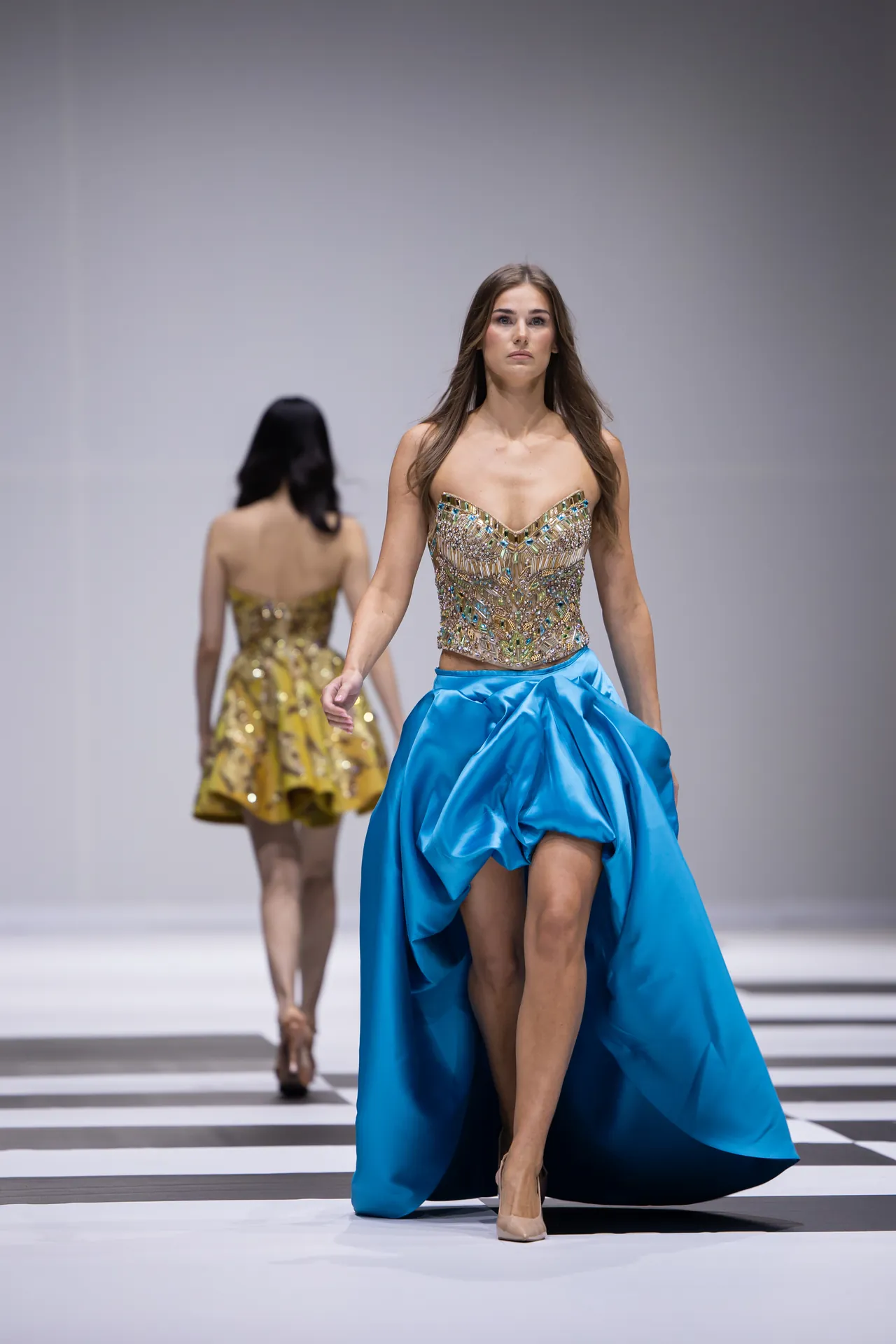 Model walking a fashion runway in a bright blue, high-low gown with an ornate jeweled bodice; another model in a gold dress follows in the background.
