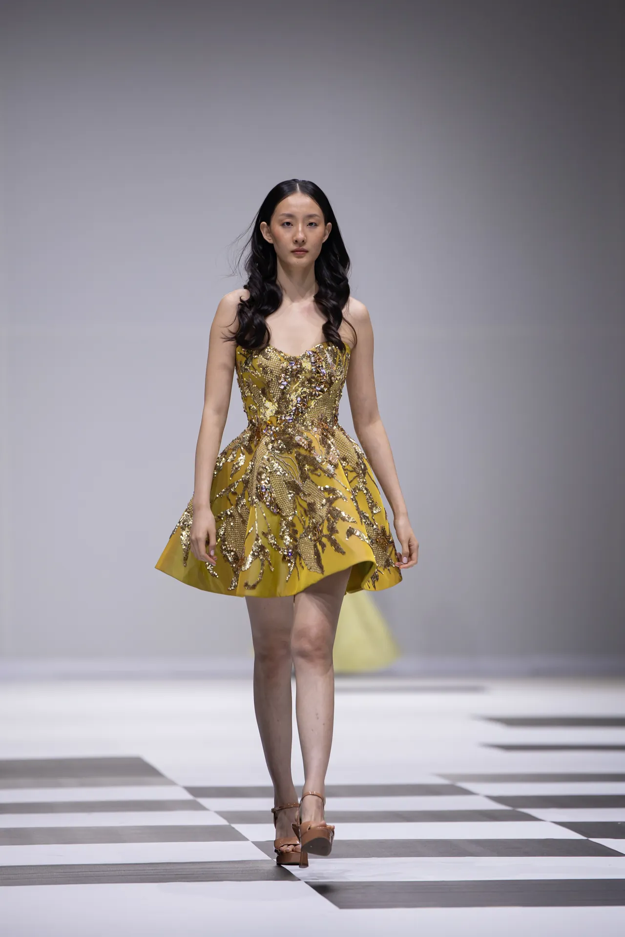 Model walking a runway in a gold and yellow embellished strapless dress with a fitted bodice and flared skirt, paired with nude heels.