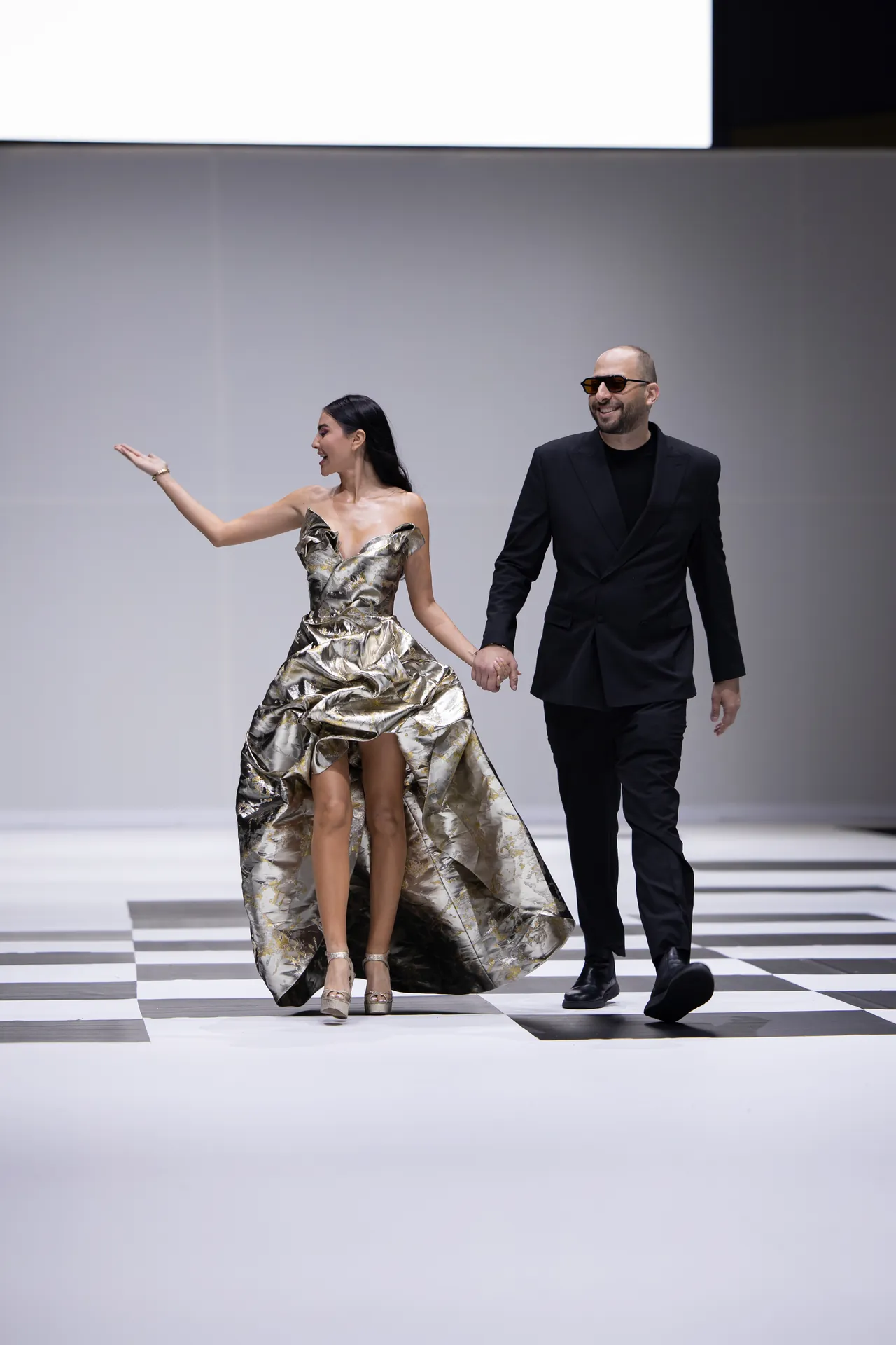 A fashionable couple walks hand in hand down a runway, the woman in a metallic strapless gown and the man in a black suit wearing sunglasses.