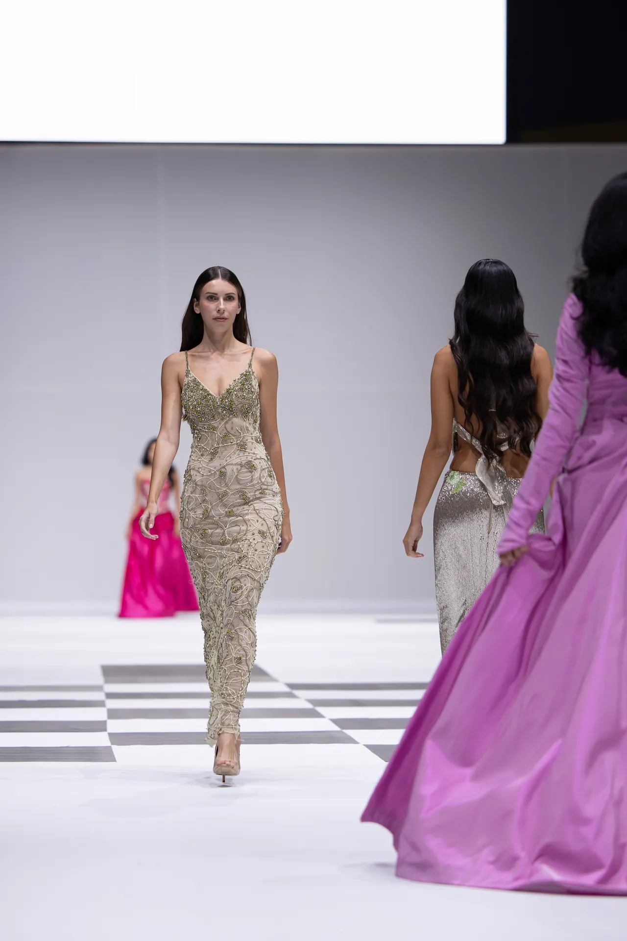 Model walks down a fashion runway in a beige embellished gown with intricate beadwork, while other models in metallic and pink gowns appear in the background on a checkered catwalk.