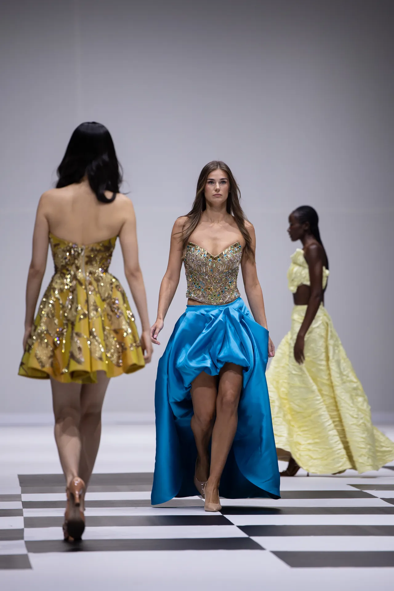 Model on a checkerboard runway wearing a strapless, bejeweled bodice with a bright blue high-low skirt; other models in gold and pale yellow gowns are visible behind her.