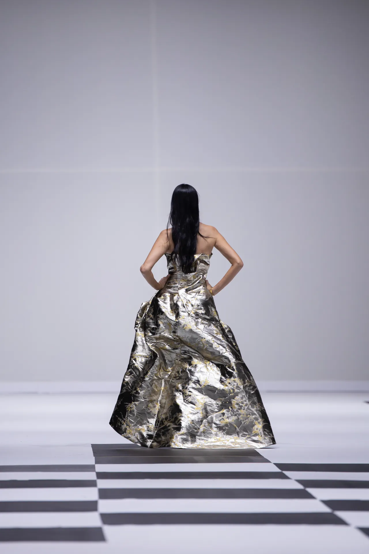 Back view of a model wearing a metallic gold gown walking on a fashion runway with a black-and-white checkered floor.