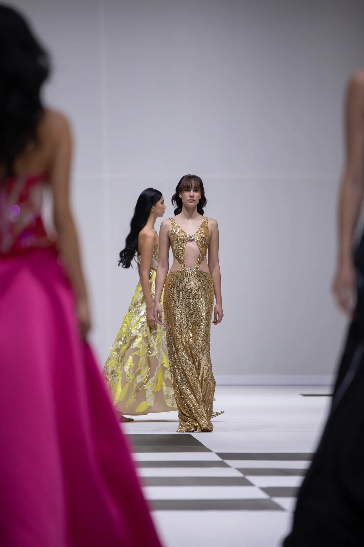 Model walks the runway in a gold sequined gown with cutouts, others in colorful gowns blurred in the background on a white checkered catwalk.