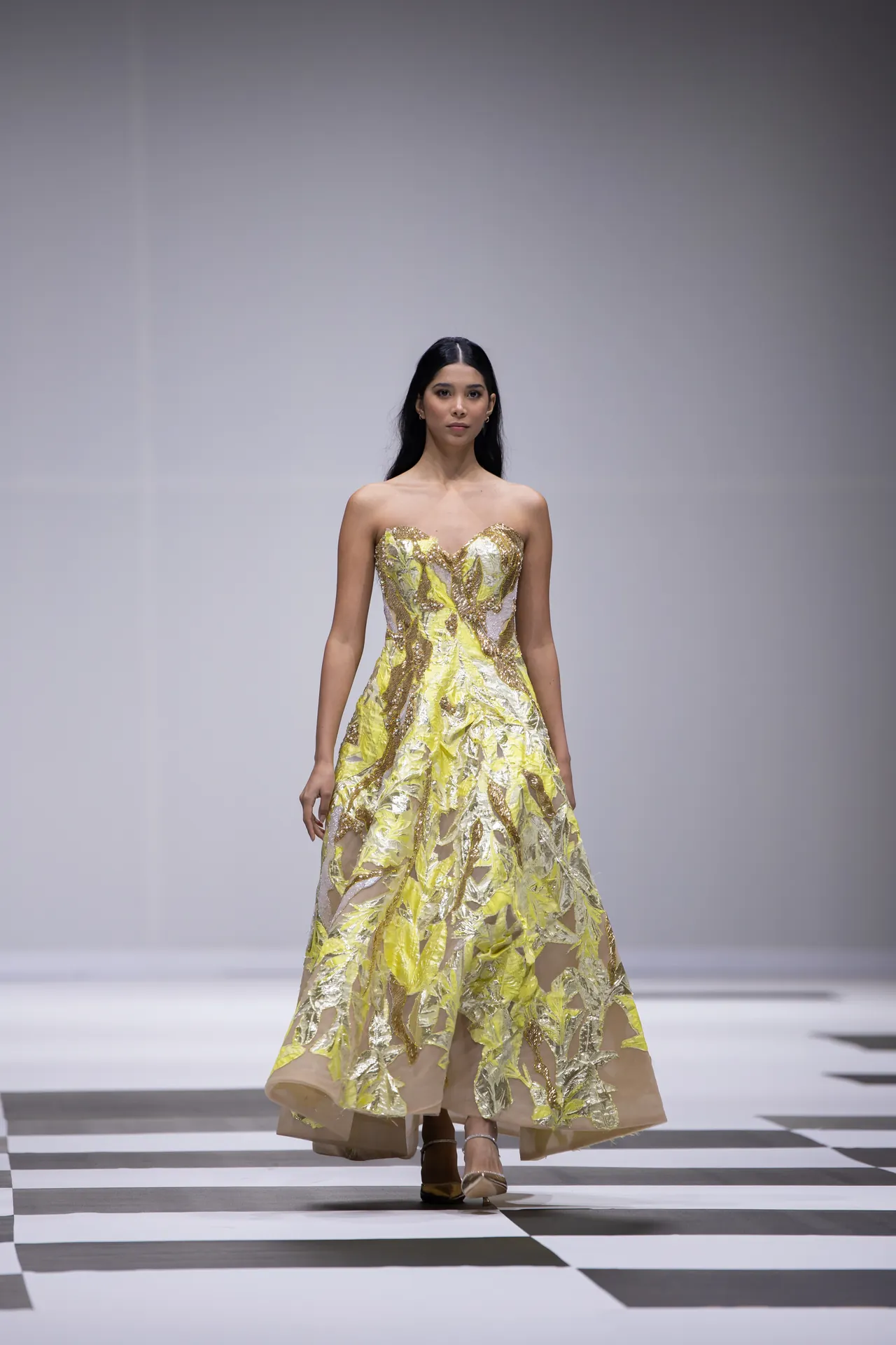 Model walking the runway in a strapless, gold-yellow embellished gown on a black-and-white checkerboard runway.
