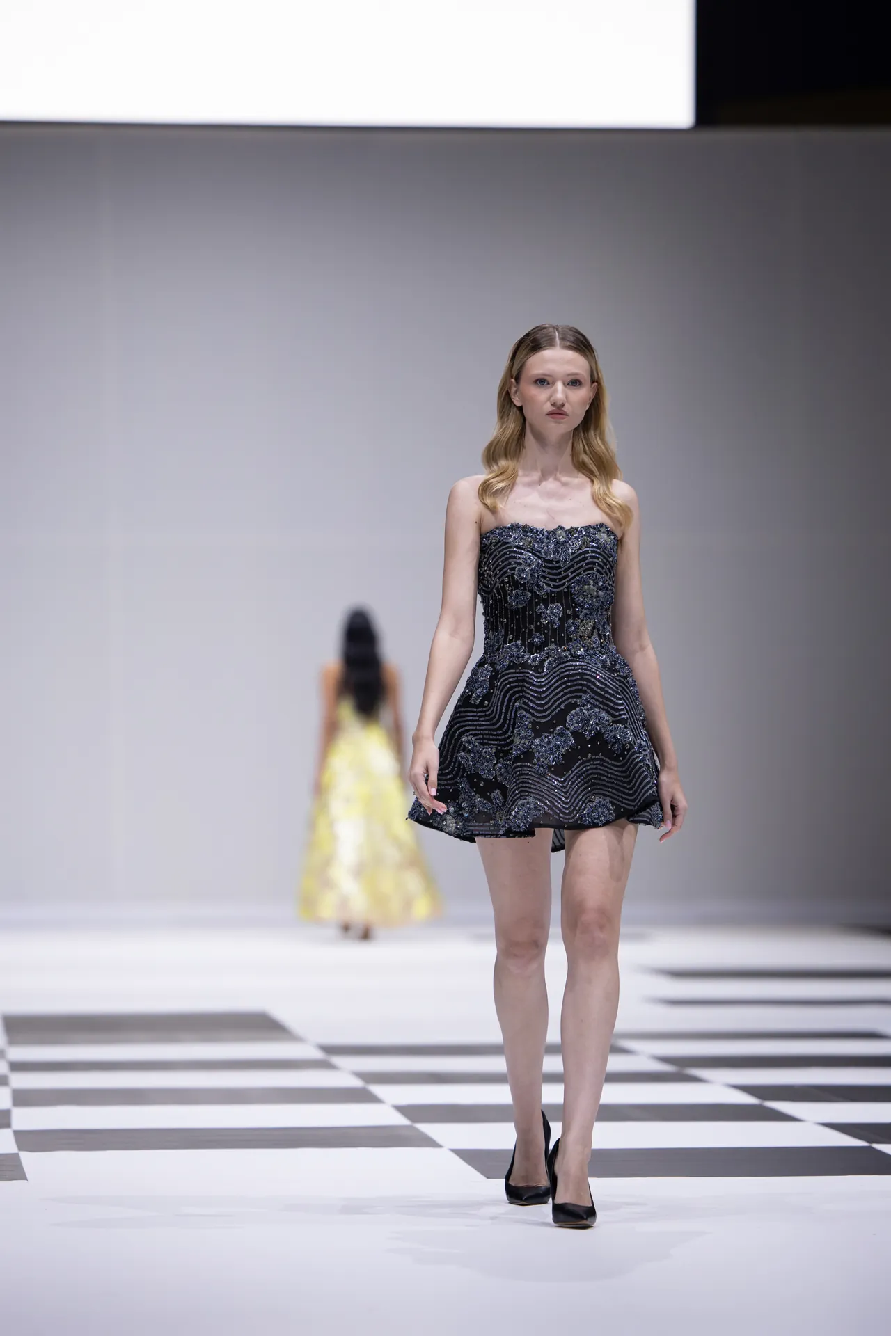 Model walks a fashion runway wearing a short, strapless black embellished dress with silver beadwork; checkered catwalk visible in foreground.