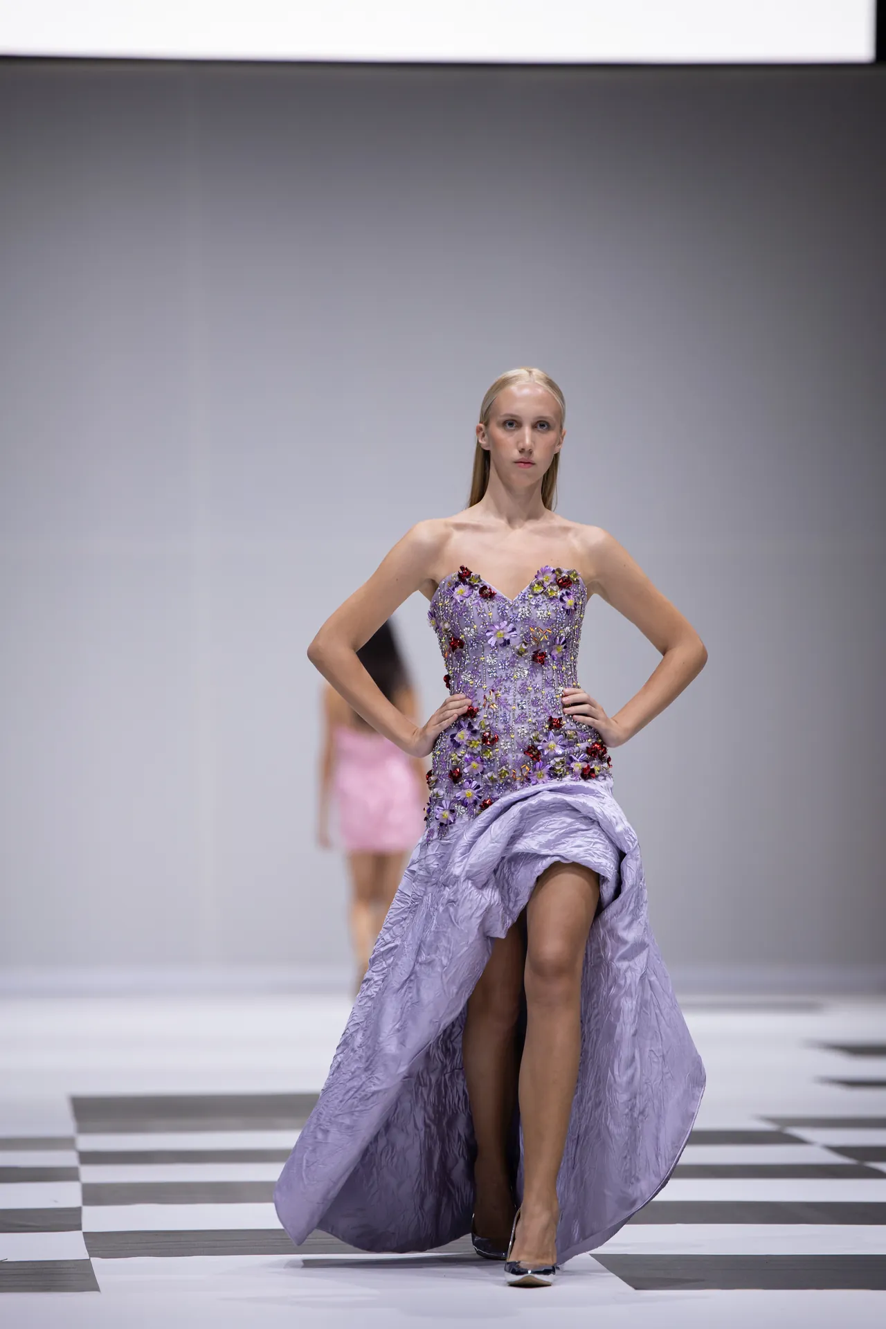Female model on a runway wearing a lavender strapless gown with floral appliques, walking down a black-and-white checkered runway.