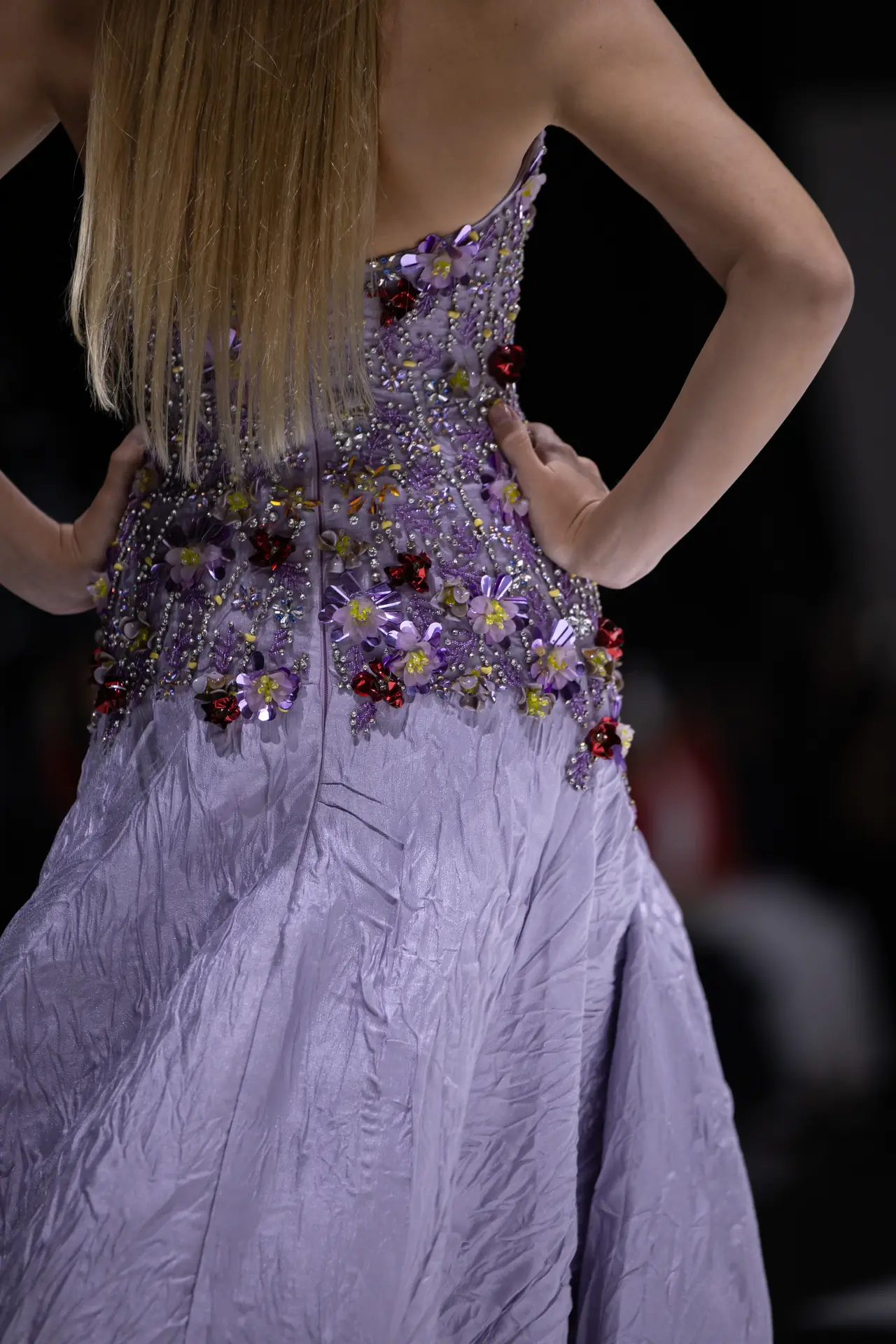 Back view of a person wearing a lilac gown adorned with purple floral appliqués and beads, facing away from the camera.