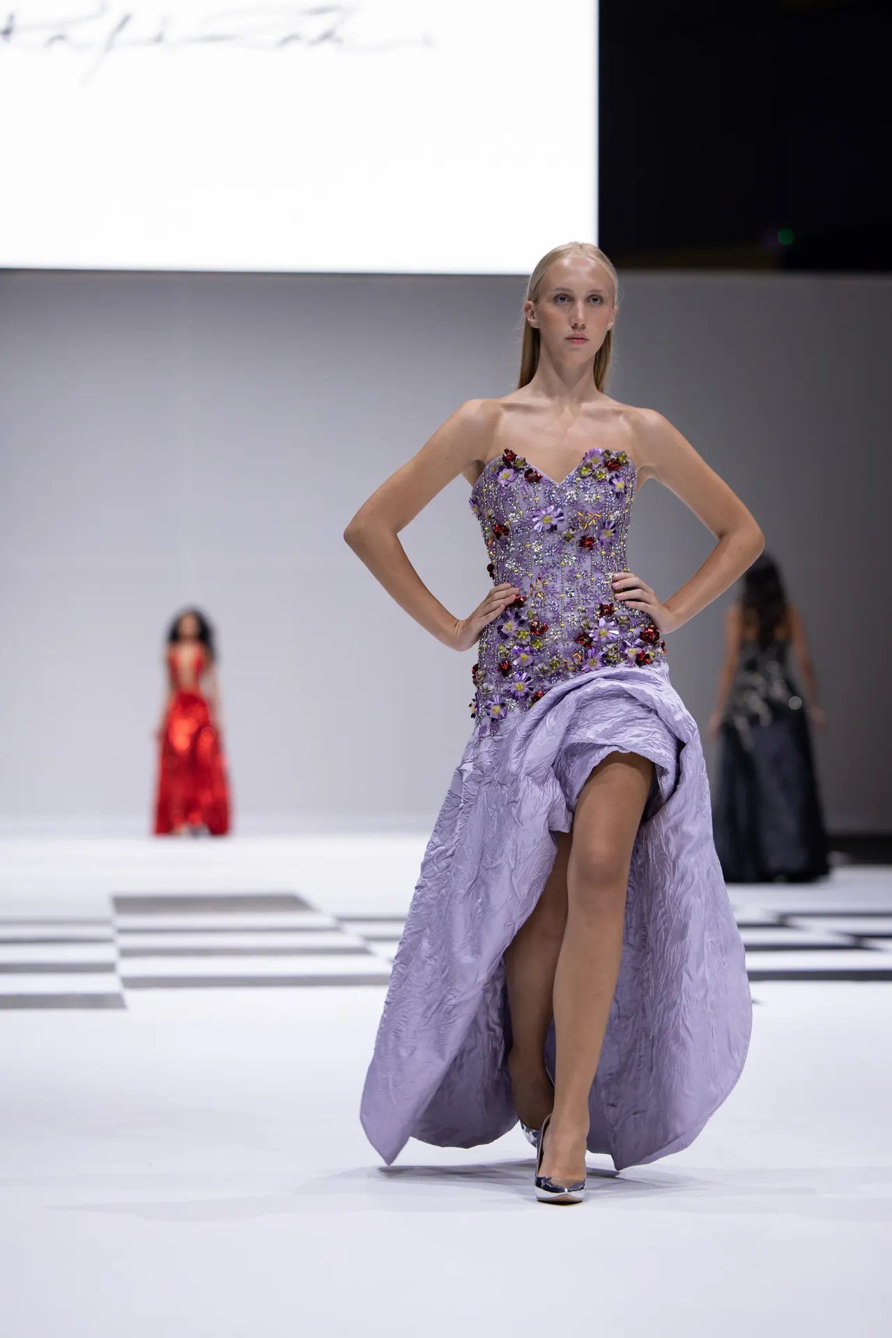 Model on a runway wearing a lavender strapless gown embellished with colorful flowers, sweeping train flowing behind her, as she poses confidently.