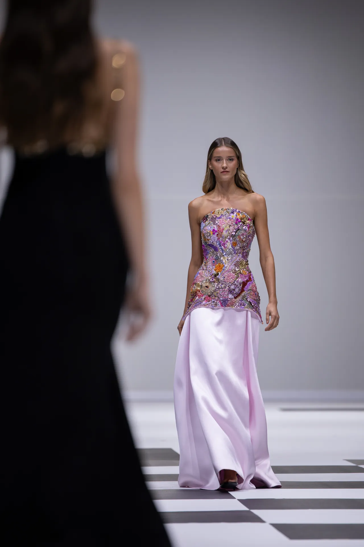 Model walks a runway in a strapless, floral-embellished gown with a pale pink satin skirt; foreground is a blurred figure on the left.