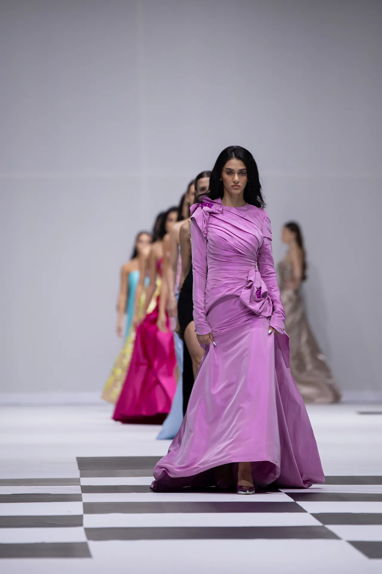Model in a lavender satin gown walks the runway, with a line of colorful models behind on a black-and-white checkered floor, against a gray backdrop.