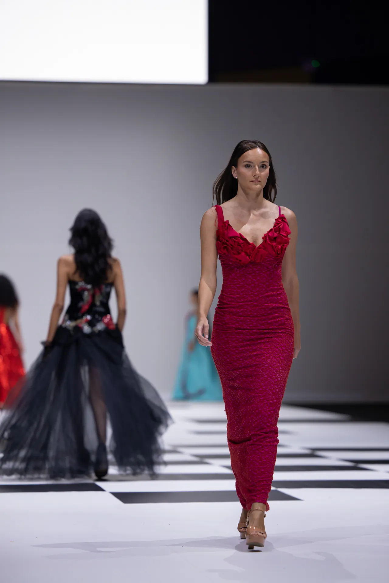 Model in a bright red lace gown walks the runway, with other models in gowns visible in the background on a checkered floor.