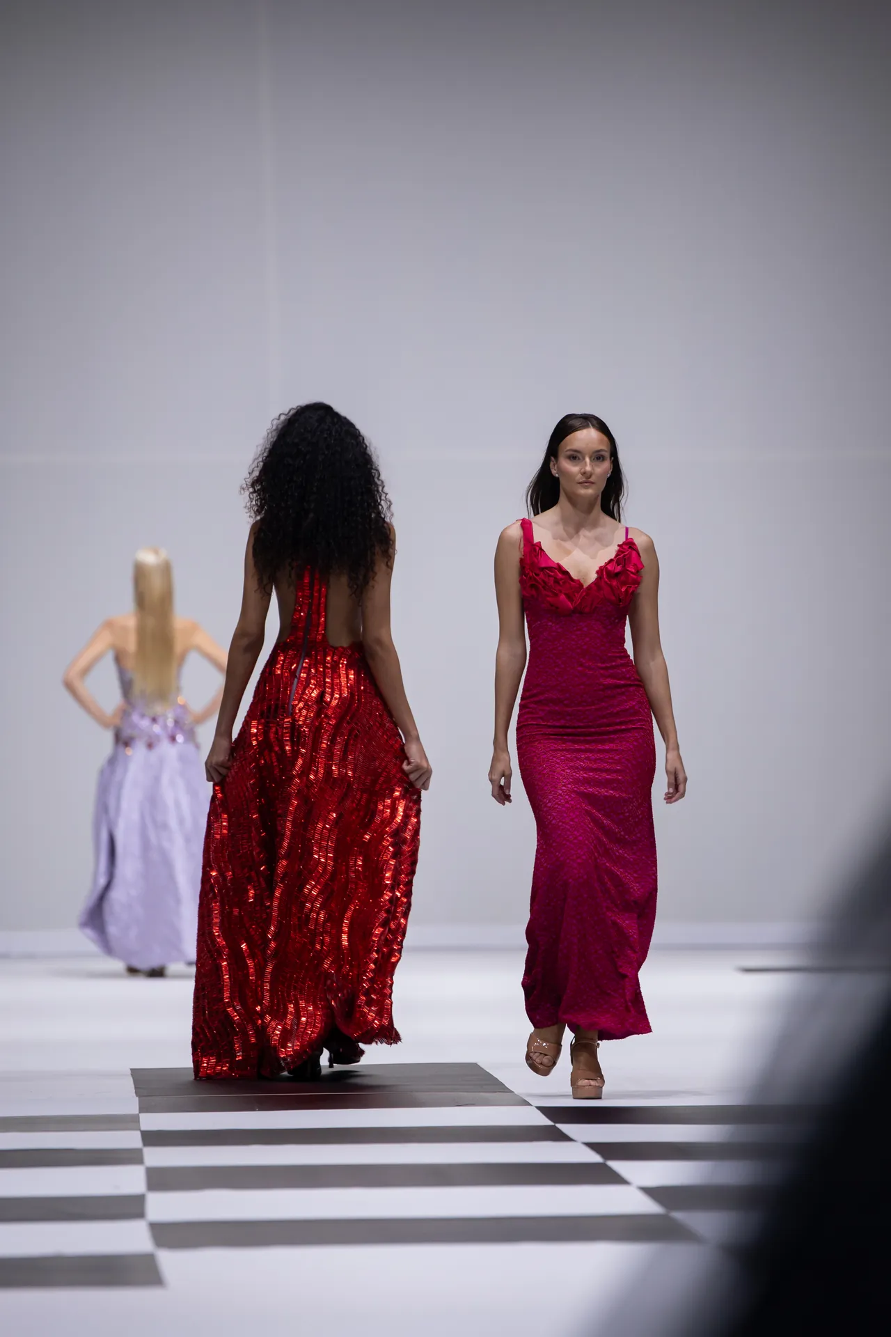 Model in a magenta lace dress walks a pink-red gown down a black-and-white checkered runway; other models pose in the background.