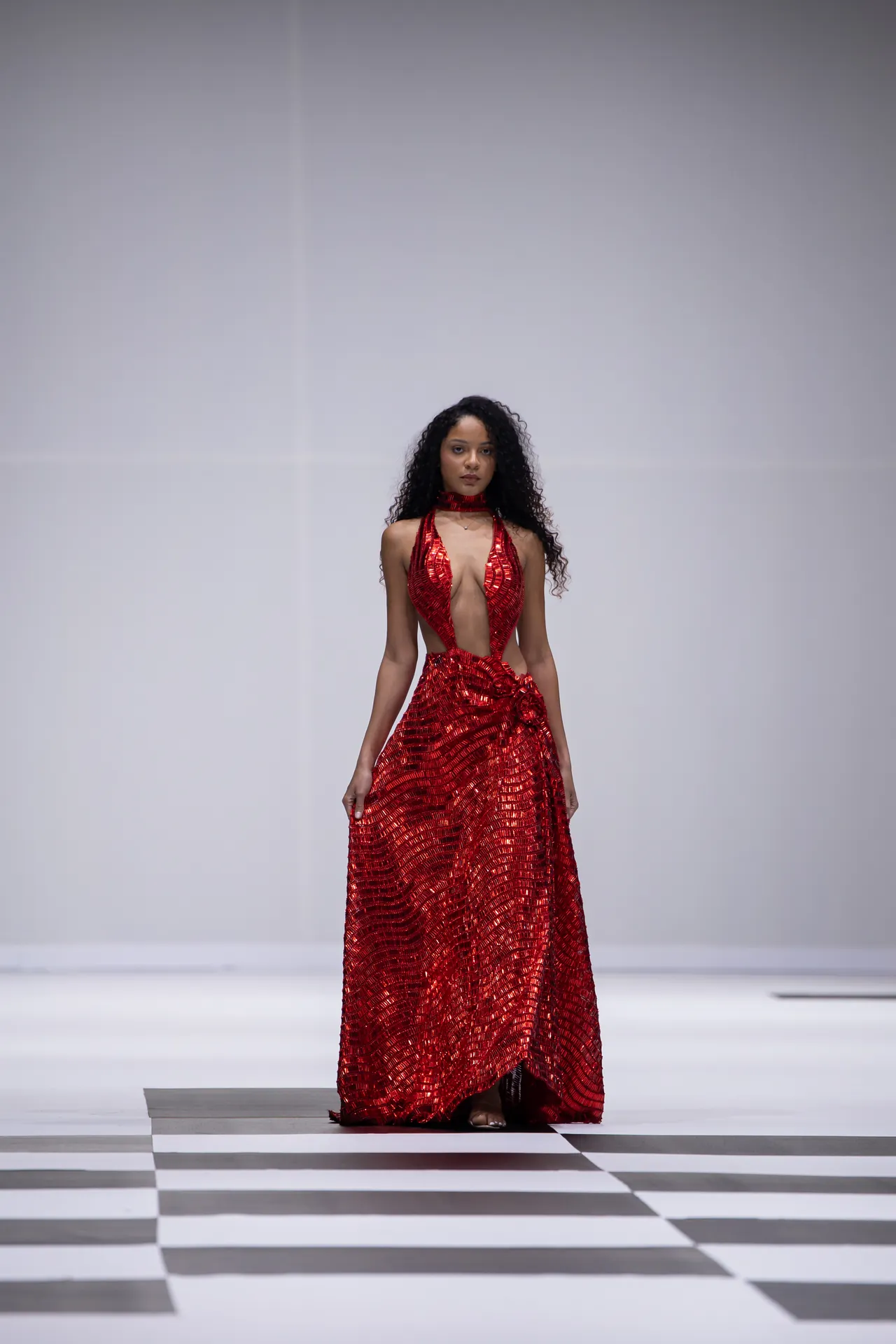 Model wearing a red sequined gown on a runway with a checkered black-and-white floor, standing centered and looking forward.”] ,