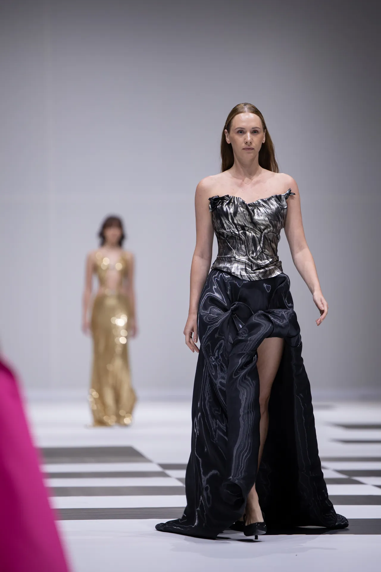 Model walks the runway in a black metallic off-shoulder gown with a high slit; another model in a gold dress is blurred in the background.