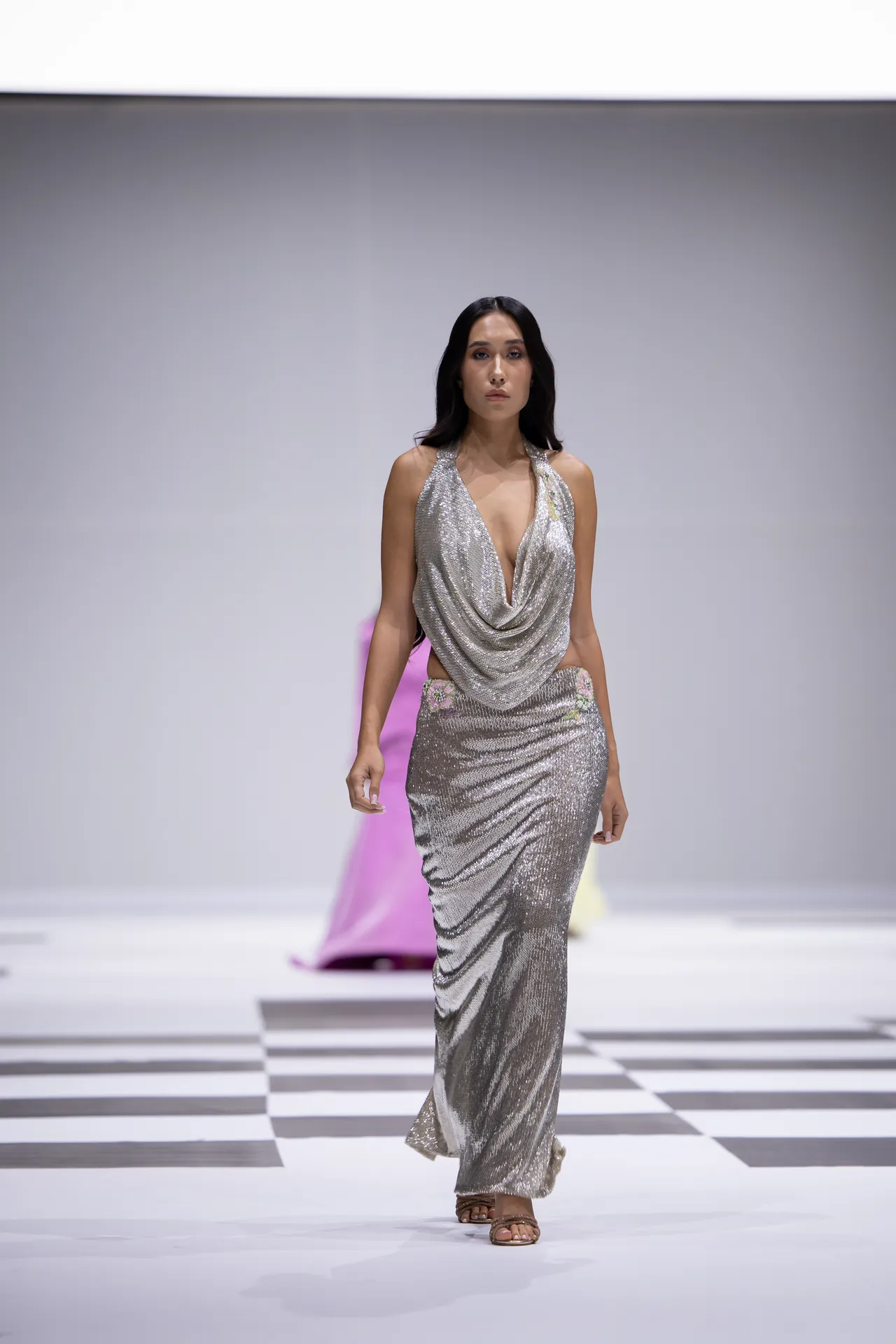 Model walking a fashion runway in a silver sequined gown with a draped neckline, mid-stride on a checkered catwalk.