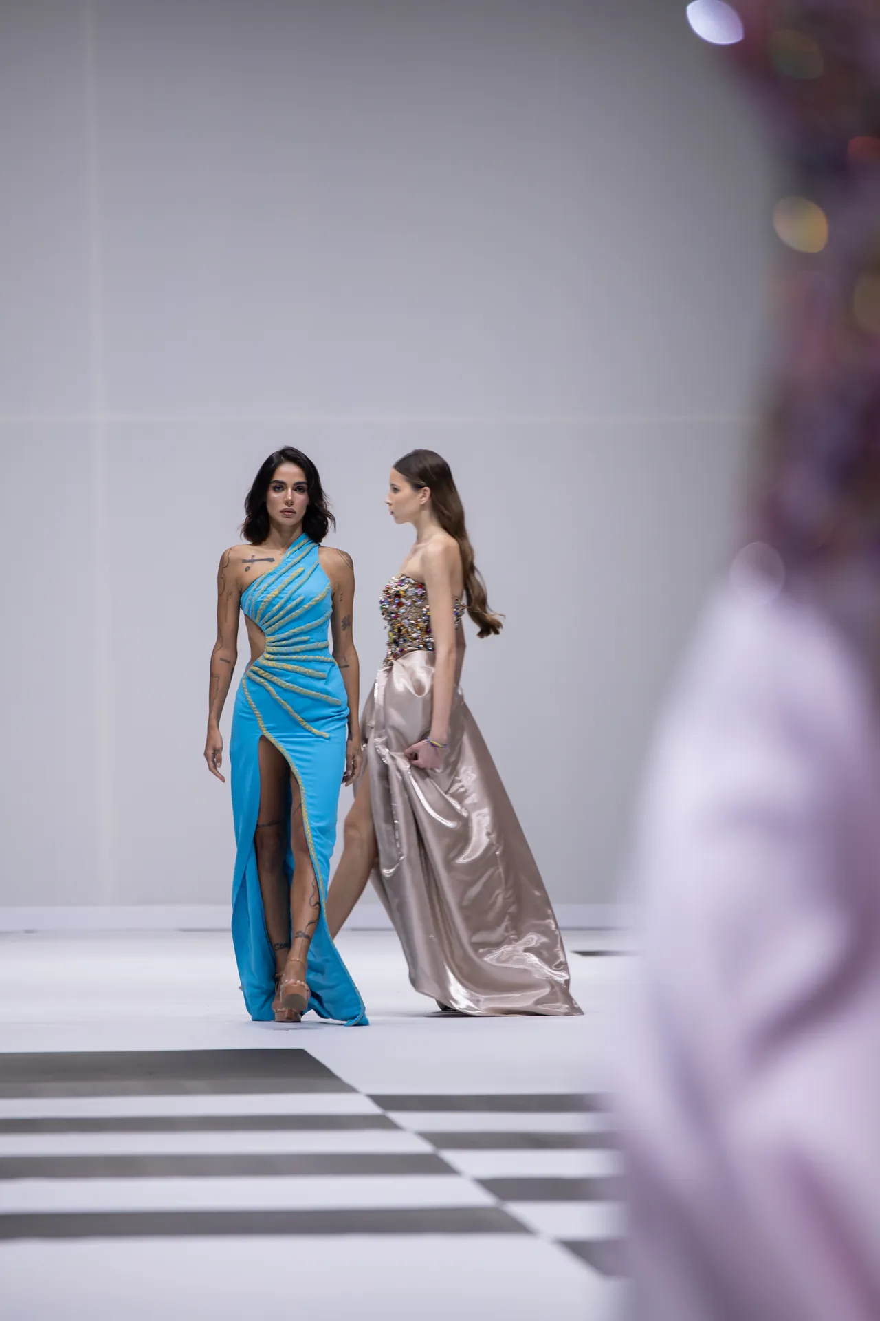 Two female models walk a fashion runway in blue and champagne gowns; blurred audience foreground on the right and a striped floor underfoot.
