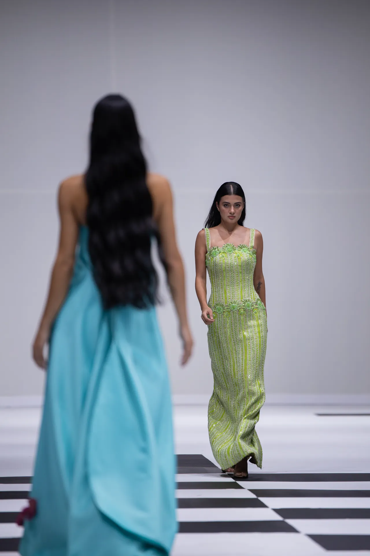 Model walks a lime green embroidered gown down a black-and-white checkered runway; foreground blurred in a turquoise dress.