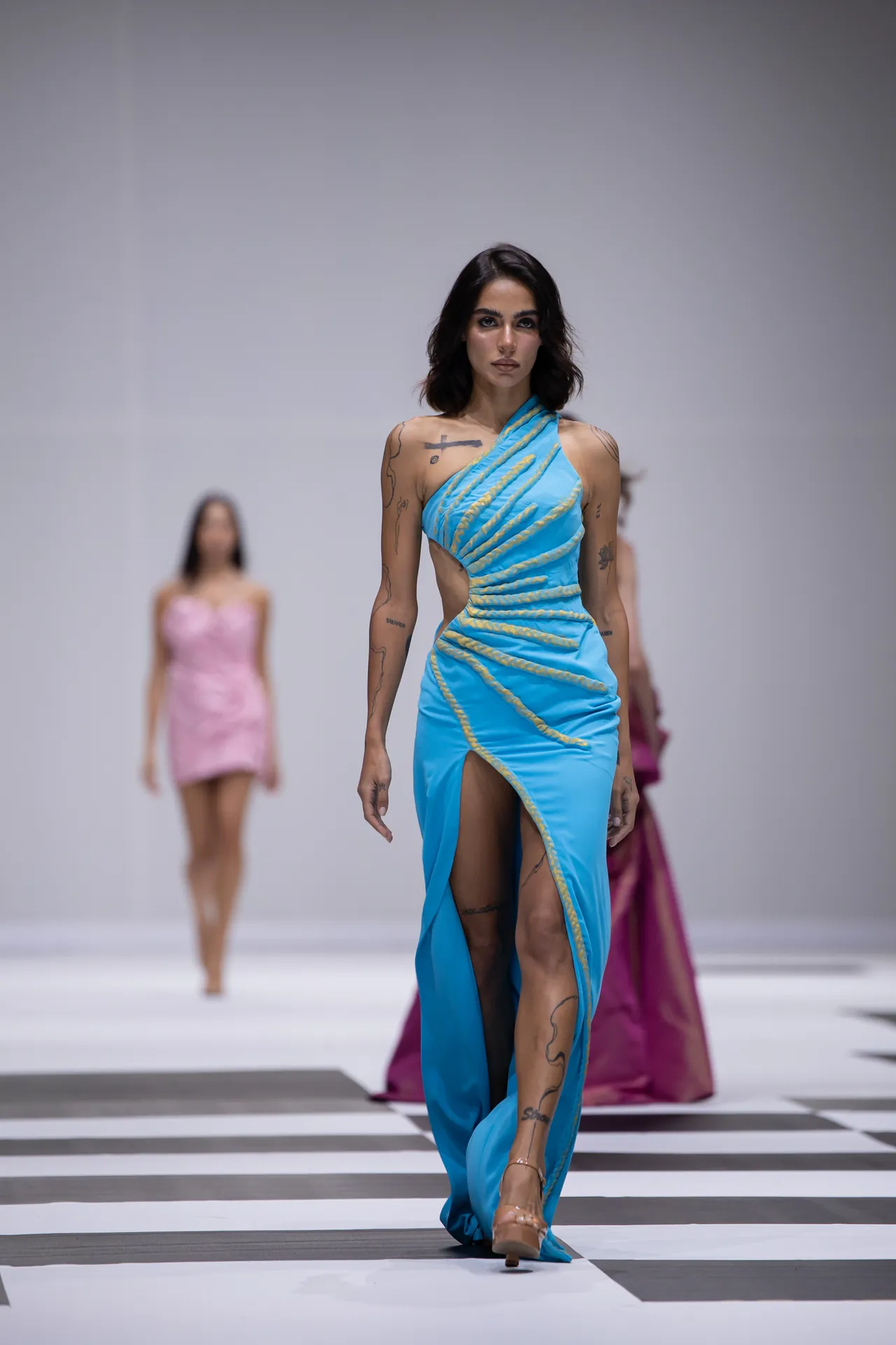 Model walking a fashion runway in a blue one-shoulder gown with gold rope accents; blurred background models and a black-and-white striped floor behind her.