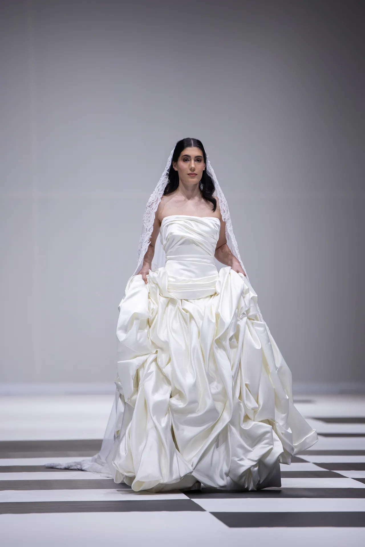 Bride in a voluminous ivory satin wedding gown with a lace veil walking a runway on a black-and-white checkered floor.