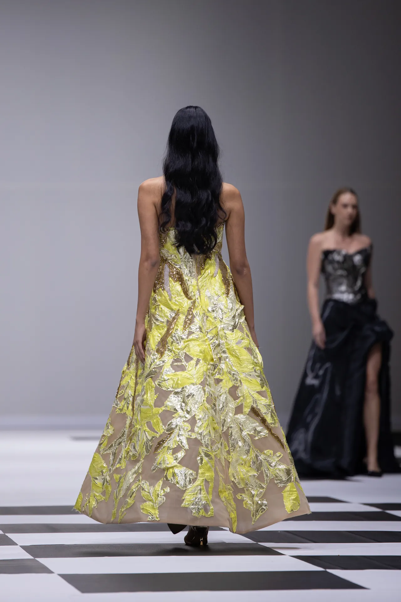 Back view of a model walking the runway in a yellow and gold leaf-embellished gown, with another model in black in the background.