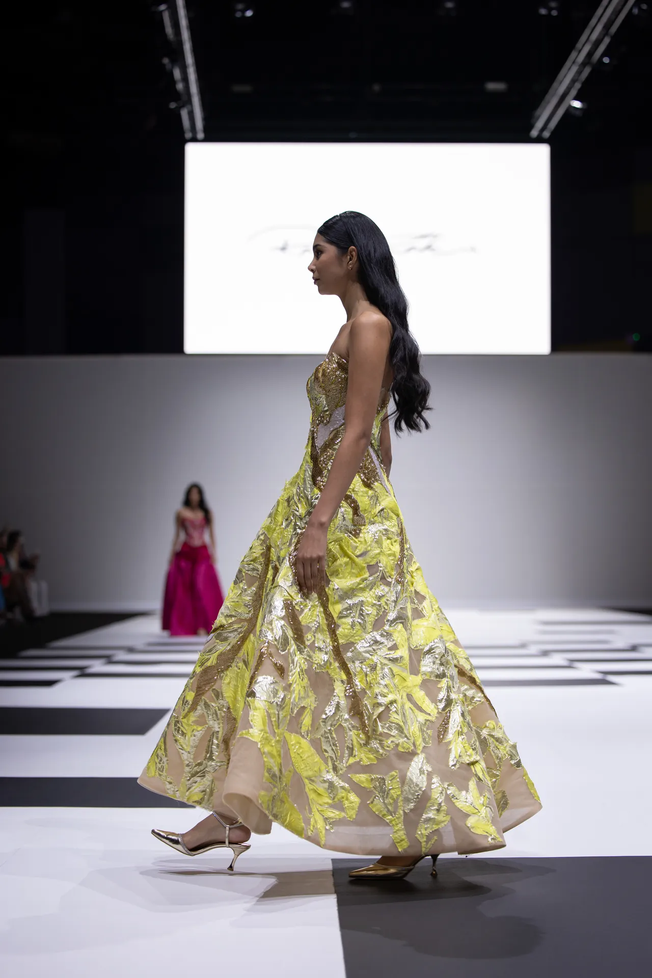 Side-profile of a fashion model walking the runway in a shimmering yellow-gold ball gown with leaf-like foil pattern on a black-and-white checkerboard catwalk.