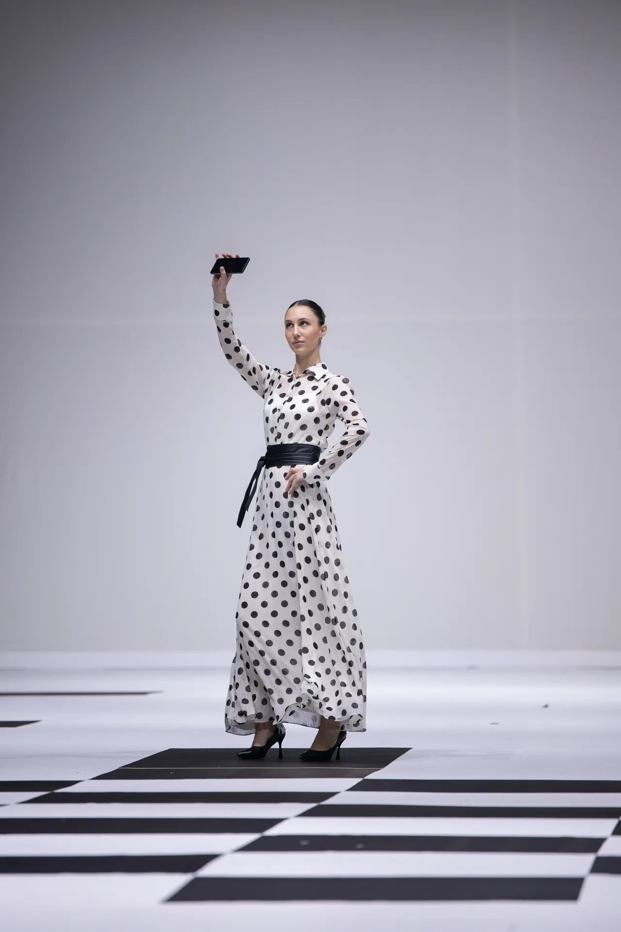 Model in a white polka-dot dress on a runway raises a tablet as if taking a selfie.