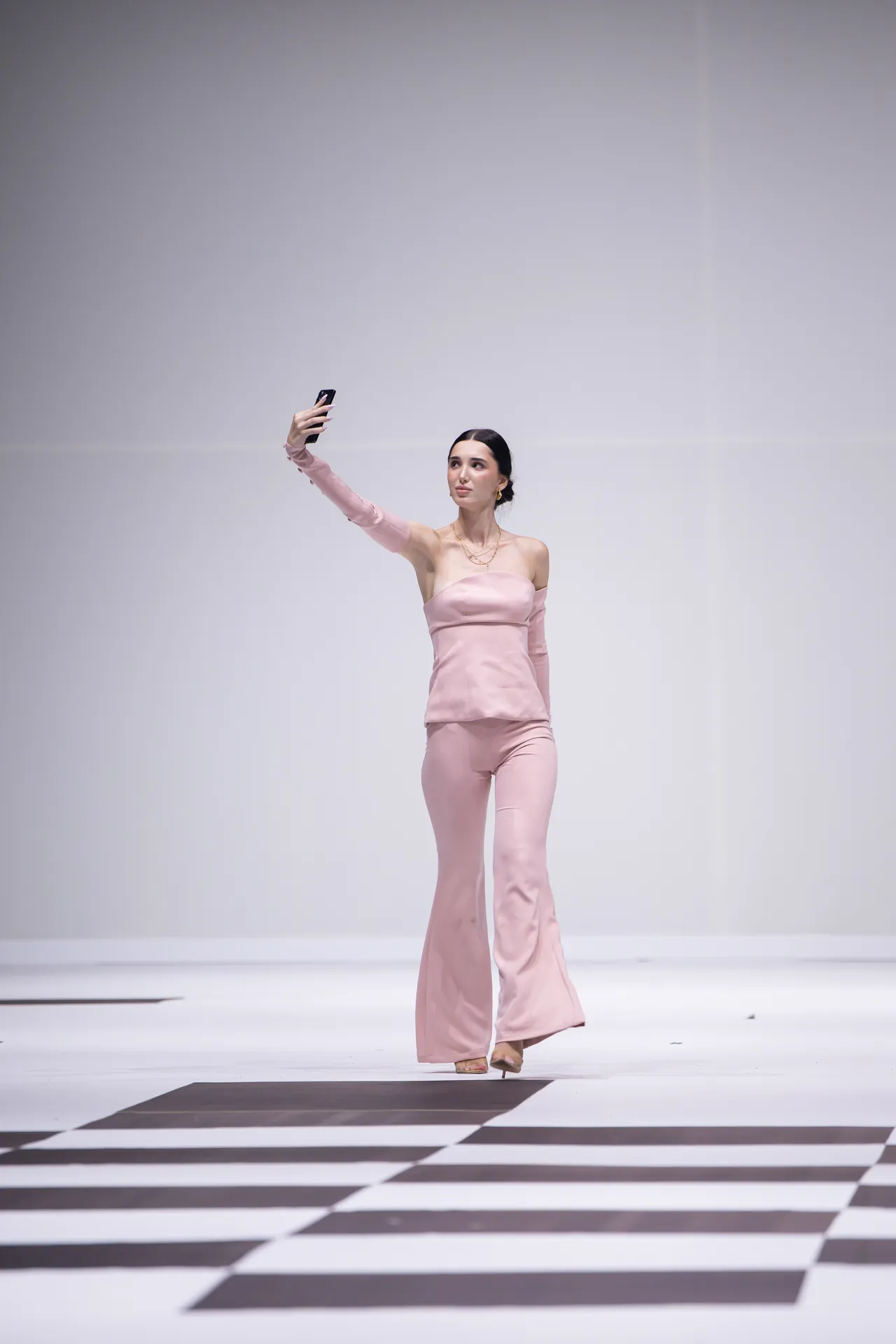 Model in a blush-pink off-shoulder top and flared pants taking a selfie on a black-and-white checker runway tile.