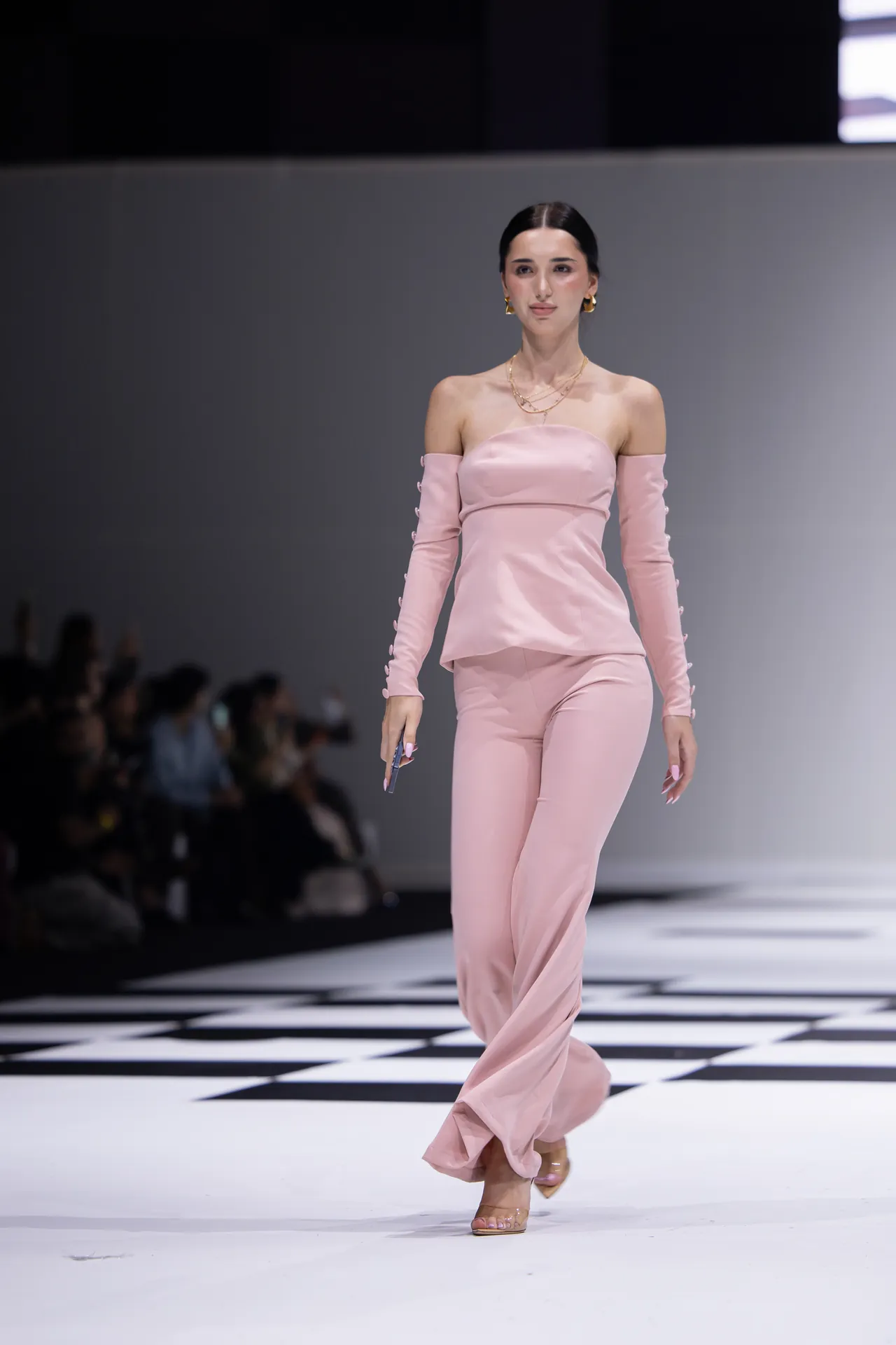 Runway model walks in a blush pink off-the-shoulder jumpsuit with long sleeves and gold jewelry, on a black-and-white checkered catwalk.