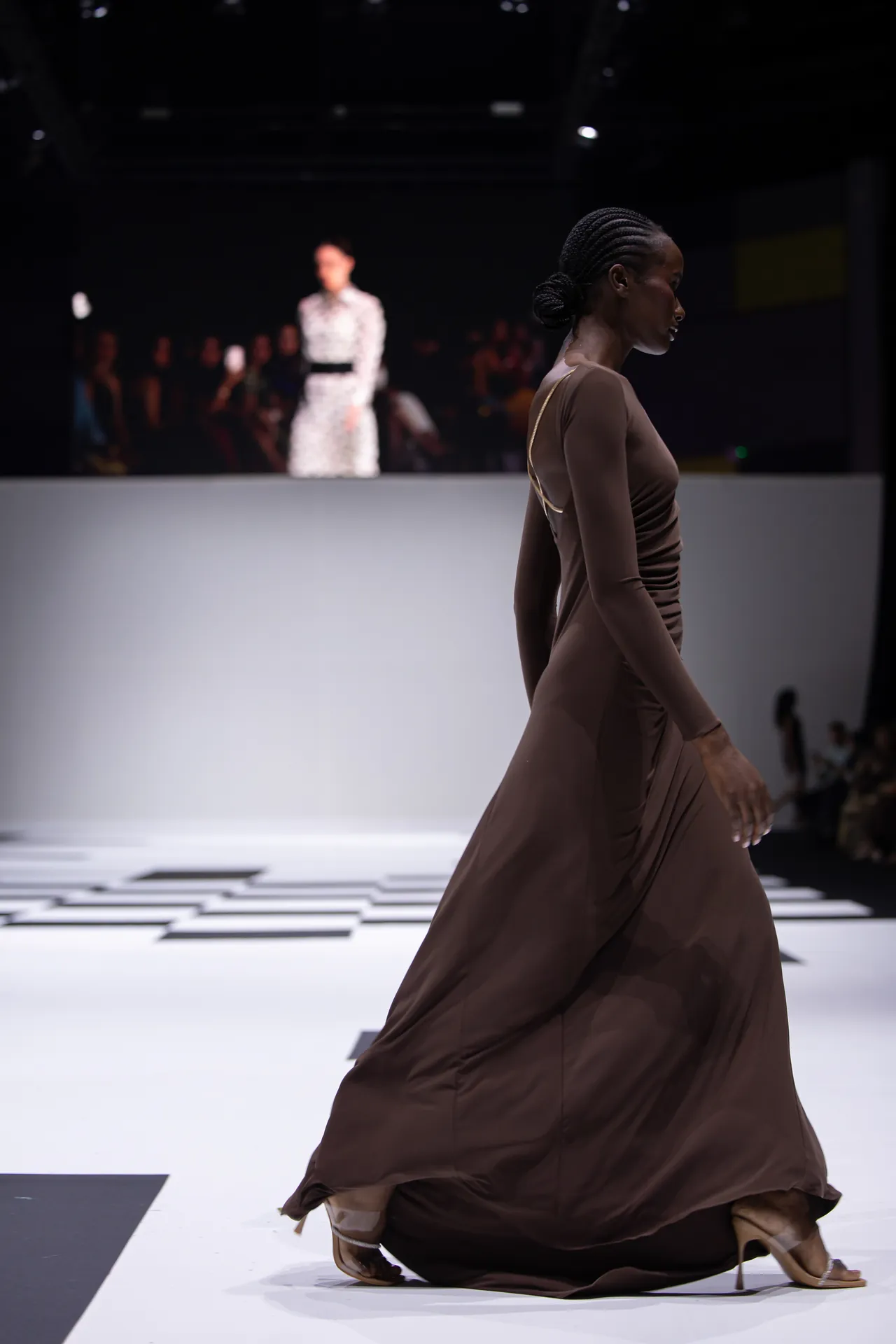 Side profile of a model in a long brown gown walking the white runway, audience blurred in the background.