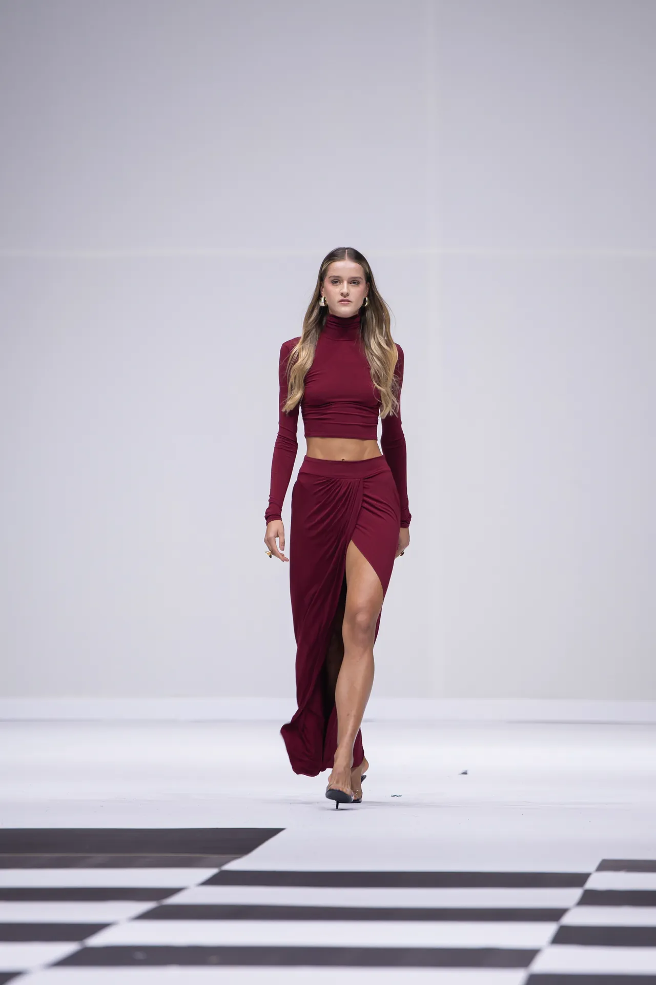 Model walks the runway in a burgundy long‑sleeve crop top and matching high‑slit skirt on a white catwalk.