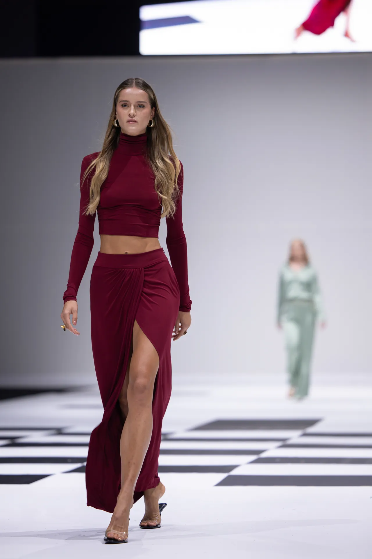 Model walking on a runway in a burgundy two-piece outfit—a turtleneck top and high-slit skirt—on a black-and-white checkered floor with a blurred model in the background.