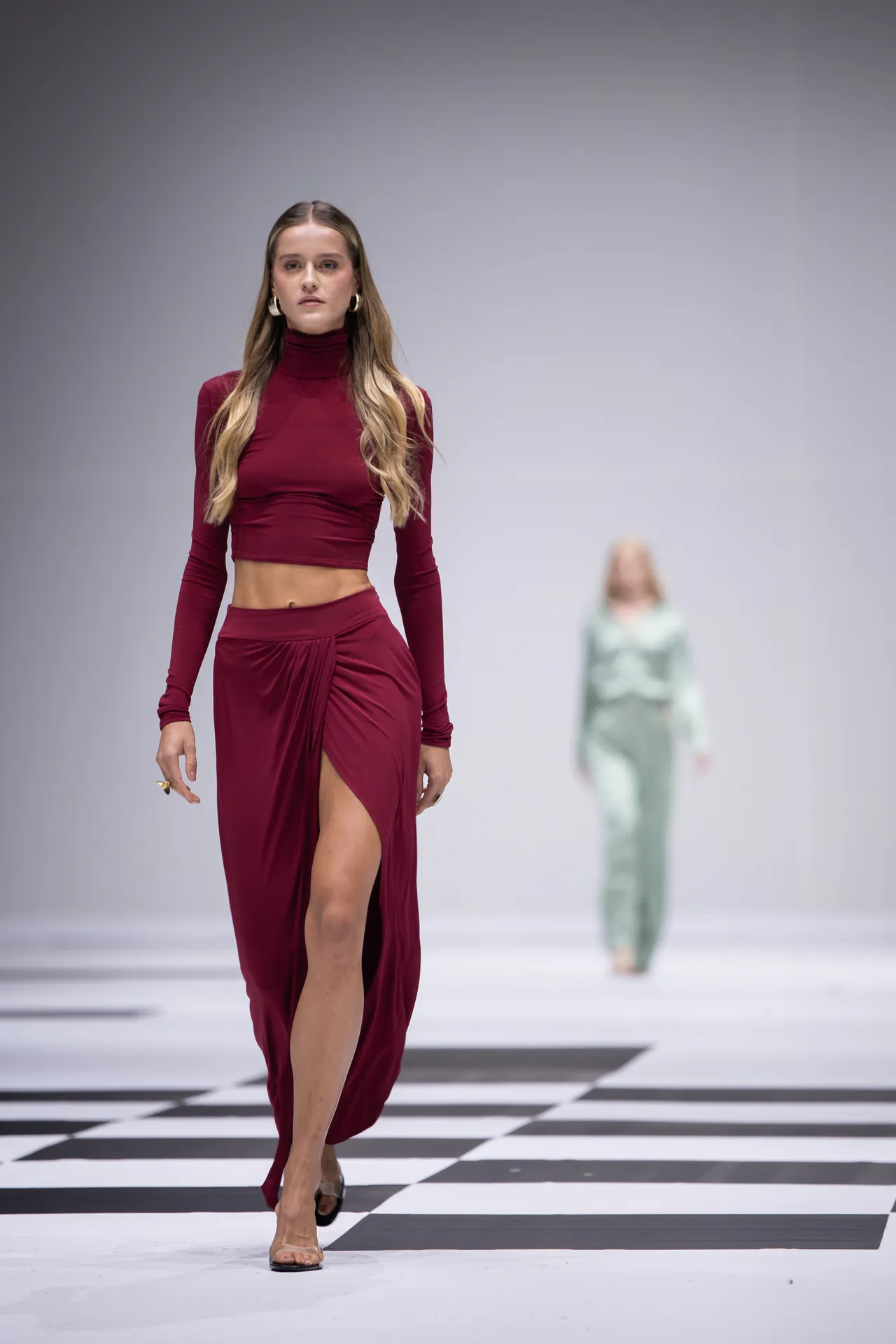 Model walks a maroon two-piece outfit (turtleneck crop top and high-slit skirt) down a black-and-white checkered runway with a blurred second model in mint green in the background.