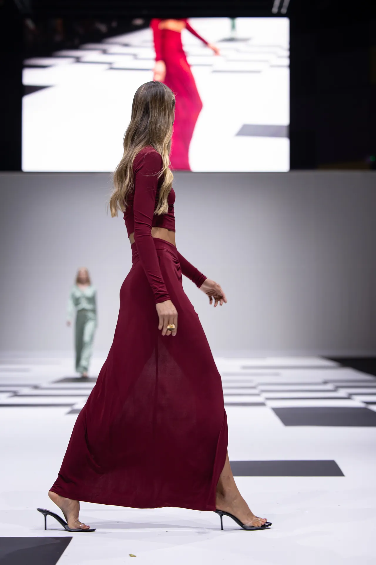 Model walking a fashion runway in a burgundy crop top and flowing skirt, wearing black heeled sandals, with a bold screen behind.