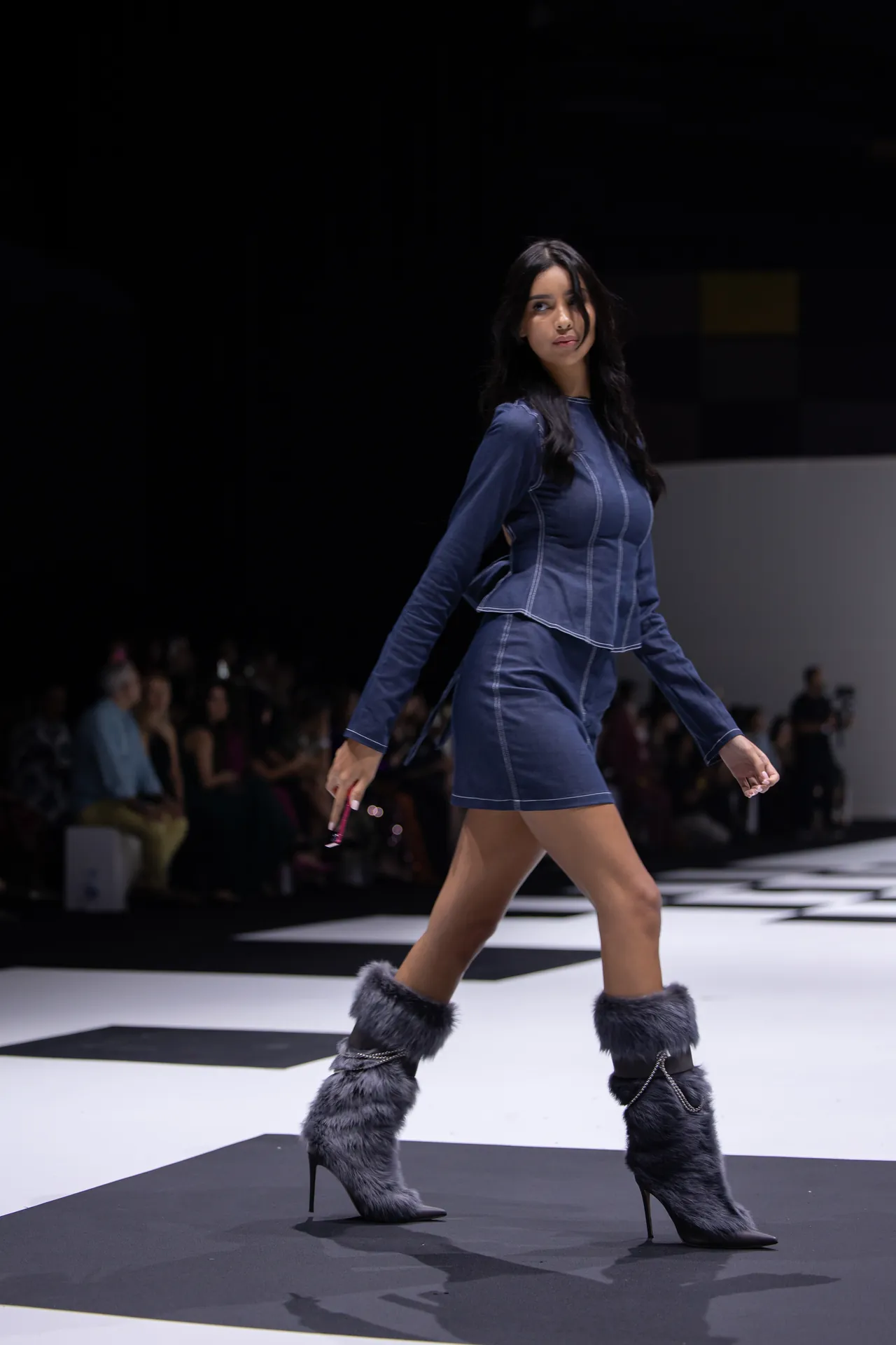 Model walks the runway in a blue, stitched dress with matching blue jacket, and tall gray furry boots; audience visible in the background.