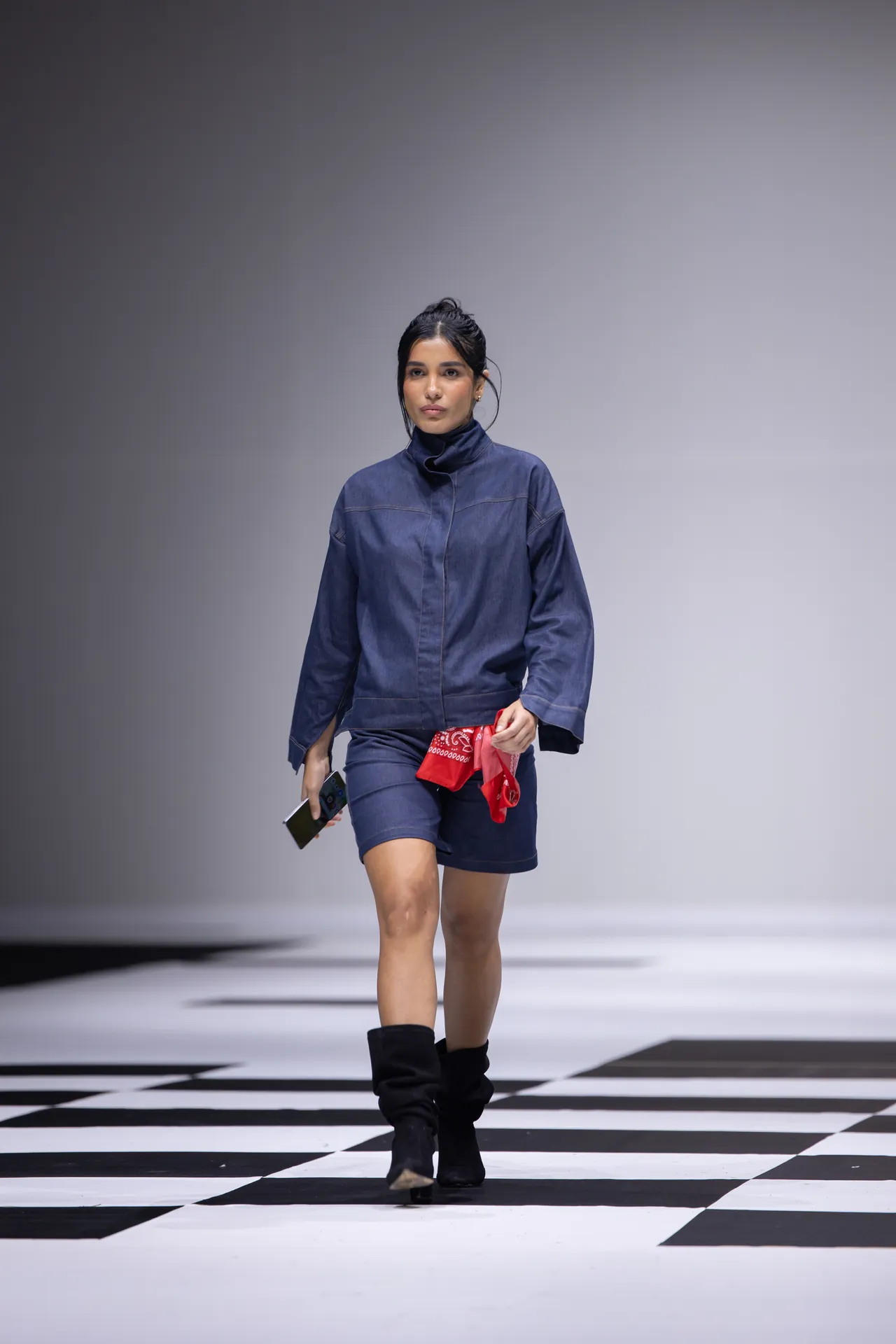 Model walking a fashion runway in a navy denim jacket and shorts, holding a red bandana and a smartphone, black boots, on a black-and-white checkered floor.