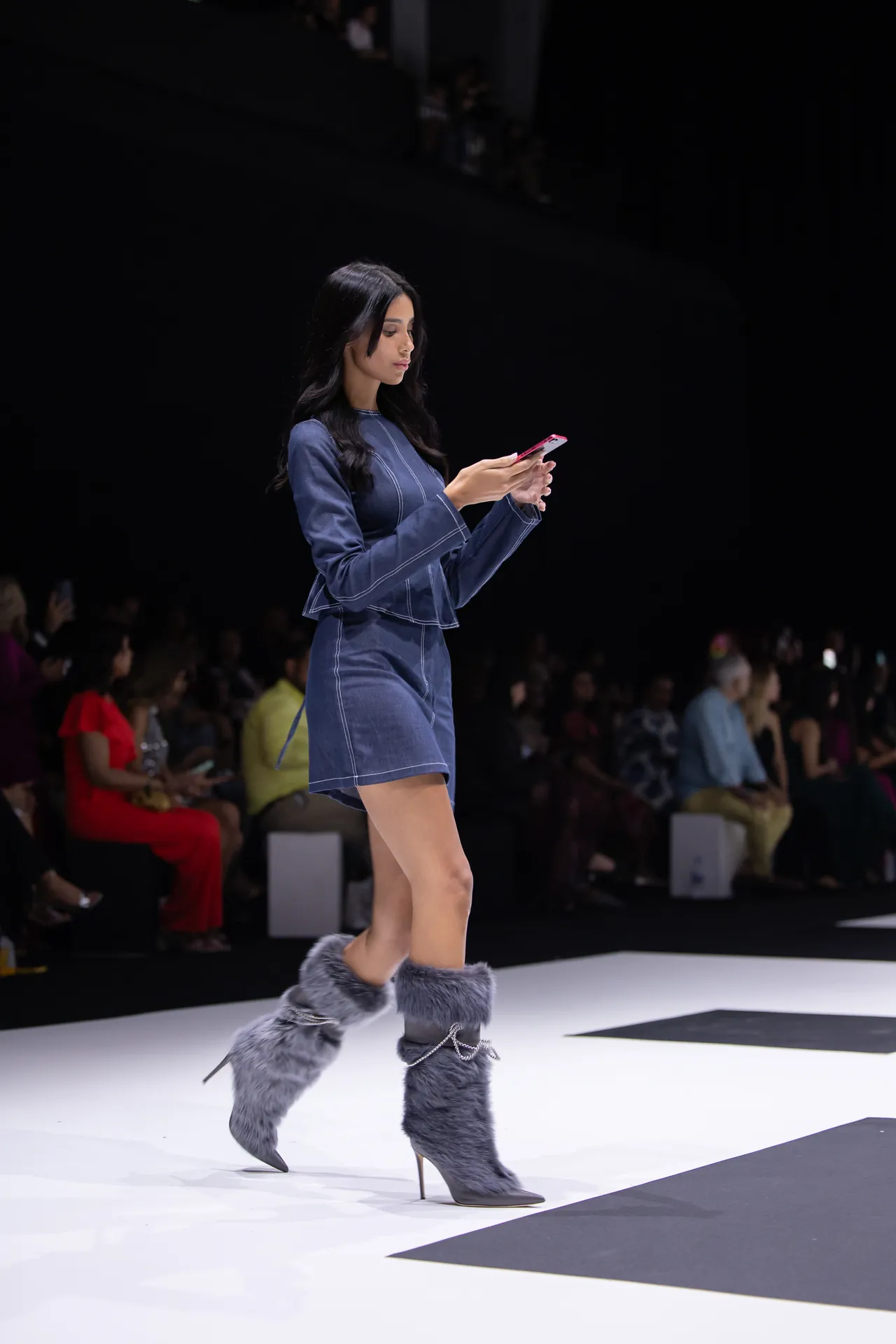 Model walking a fashion show runway in a blue denim outfit with gray fur boots, audience in the background blurred.