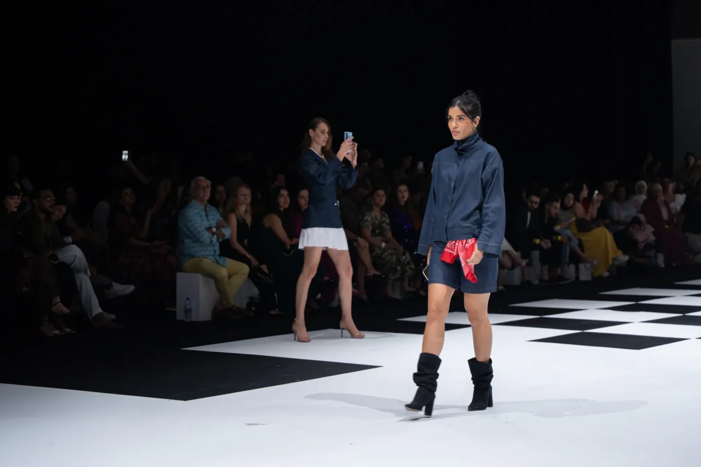 Model in a blue denim jacket and shorts walks the runway; black boots, red scarf in hand, with an audience in dim lighting behind.