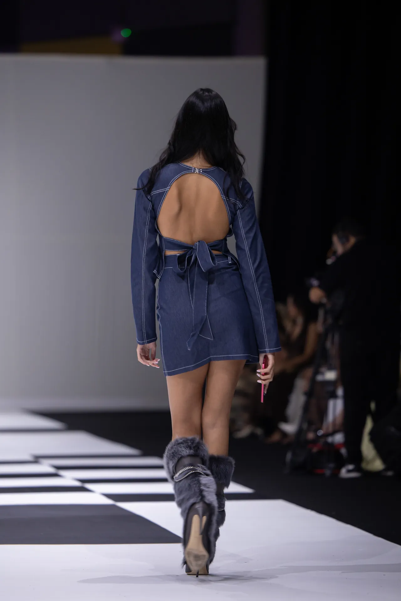Model walking away on a fashion runway in a blue denim open-back dress with a tied waist and fur-trimmed boots; audience visible in the background.