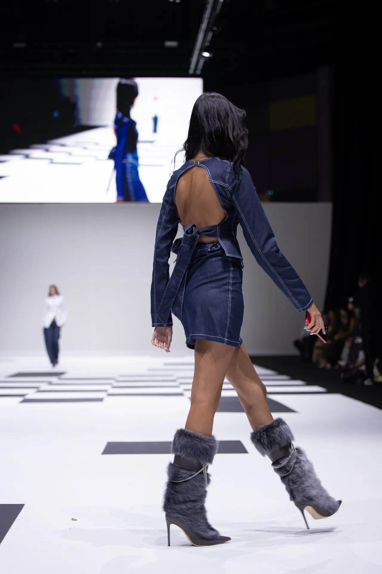Model walks the runway in a dark denim backless top and matching shorts, with gray fur-lined ankle boots on a checkered white catwalk.