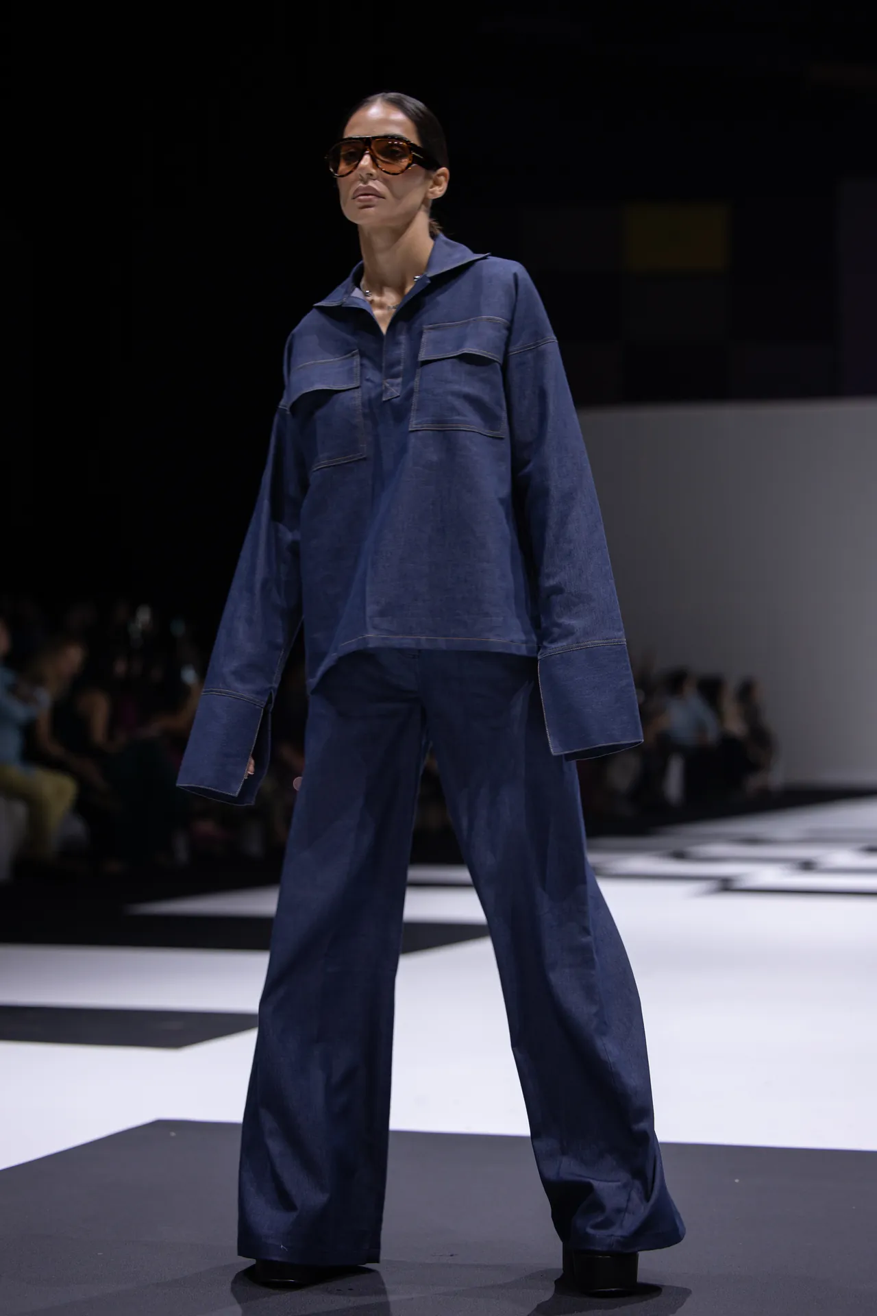 Model walking a runway in an oversized blue denim jumpsuit with large front pockets and wide legs, wearing brown sunglasses.