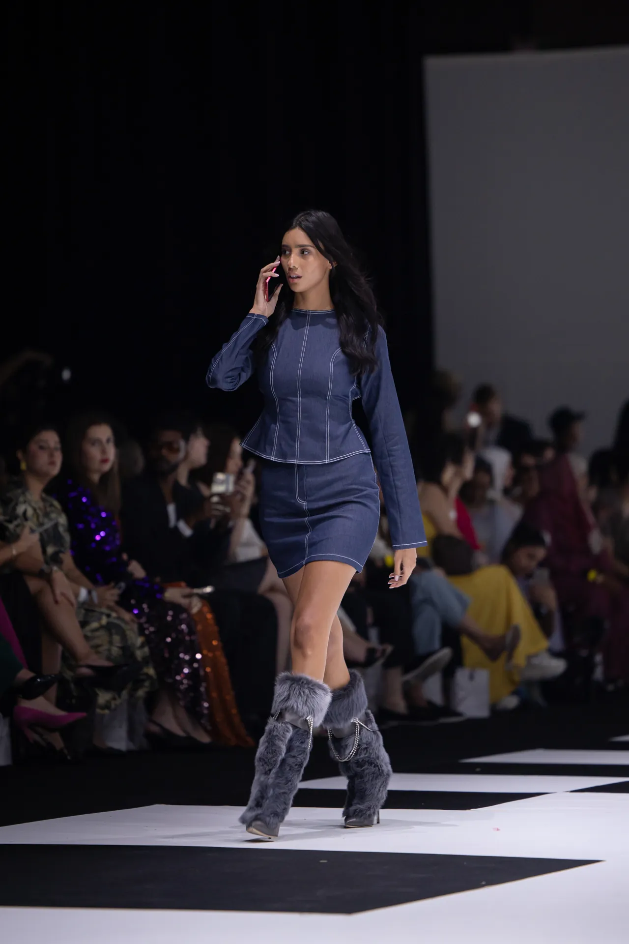 Model walks the runway in a blue denim peplum top and skirt, talking on a pink phone, audience in the background.