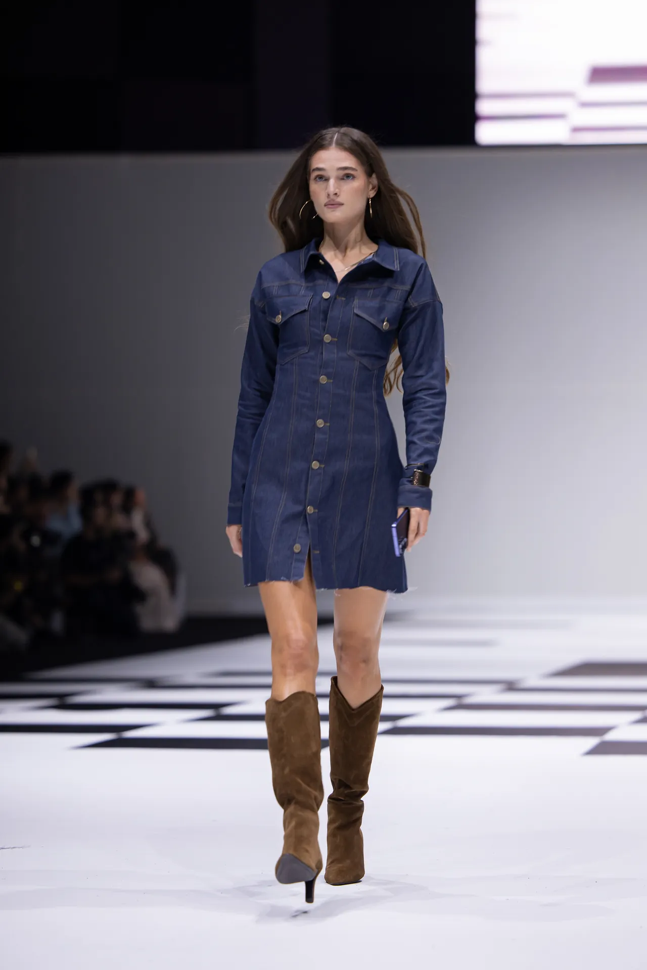 Model walks a denim shirt-dress with gold buttons on a white runway, knee-high brown boots visible from the front.